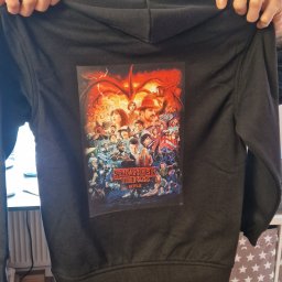 EMBE Maciej Piotrowski - Czarna bluza z kapturem, na plecach nadruk z motywem 'Stranger Things', trzymana przez osobę. Widoczne spodnie dresowe i fragment regału z pudełkami w gwiazdki.