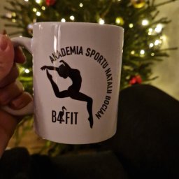 EMBE Maciej Piotrowski - Biały kubek z czarnym nadrukiem logo 'Akademia Sportu Natalii Bocian B4FIT' na tle choinki z lampkami, trzymany w dłoni. Prezentacja identyfikacji wizualnej.