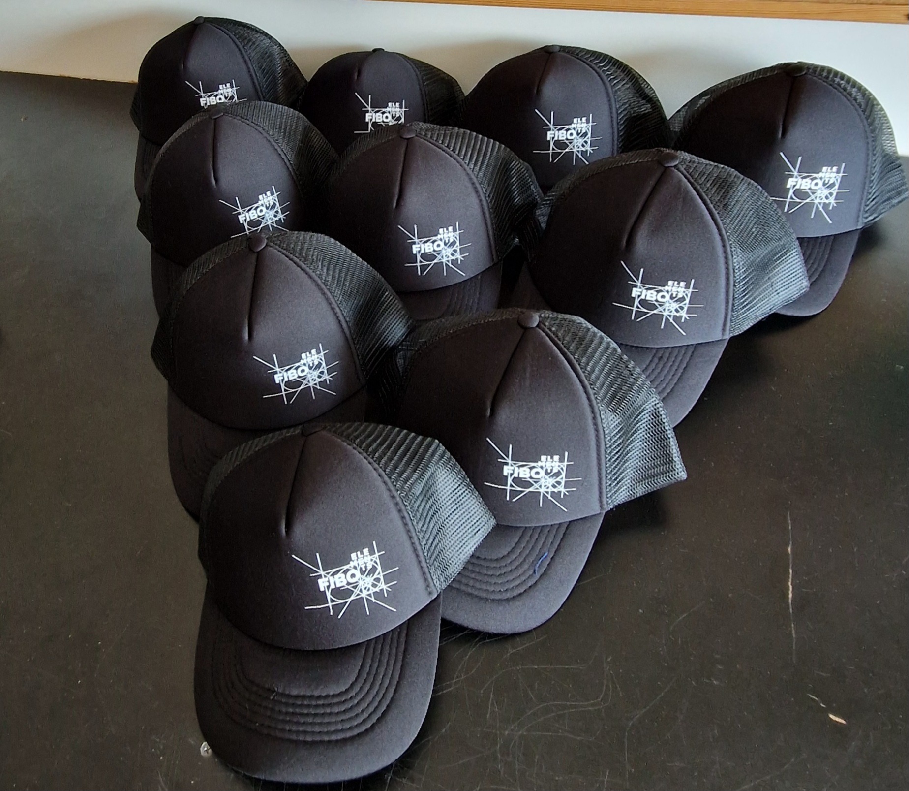 Stos czarnych czapek z daszkiem typu trucker z białym nadrukiem logo 'ELE FIBOU' na froncie, ułożonych na ciemnej powierzchni. Widoczne detale siateczki i szwów.