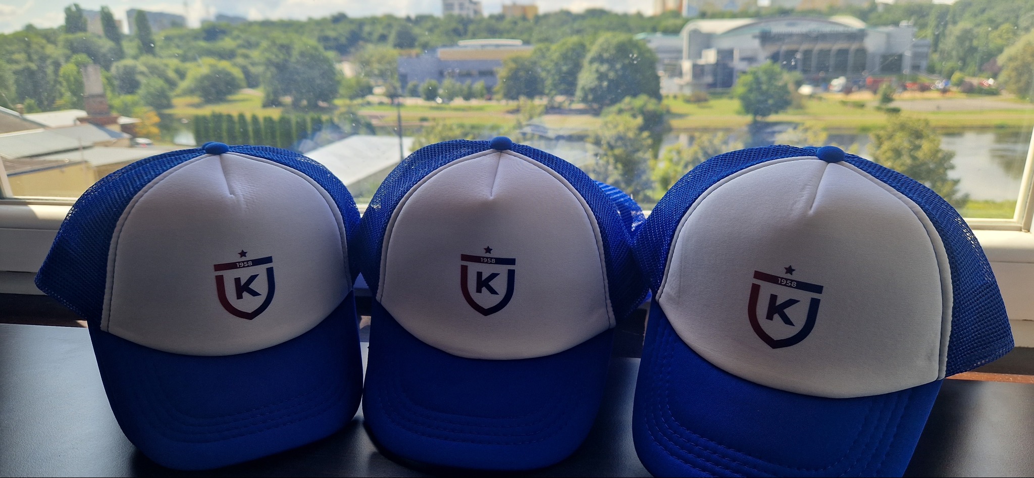 Trzy identyczne, biało-niebieskie czapki z daszkiem, z logo w kształcie tarczy z literą K i rokiem 1958, ustawione na tle okna z widokiem na zieleń miejską.