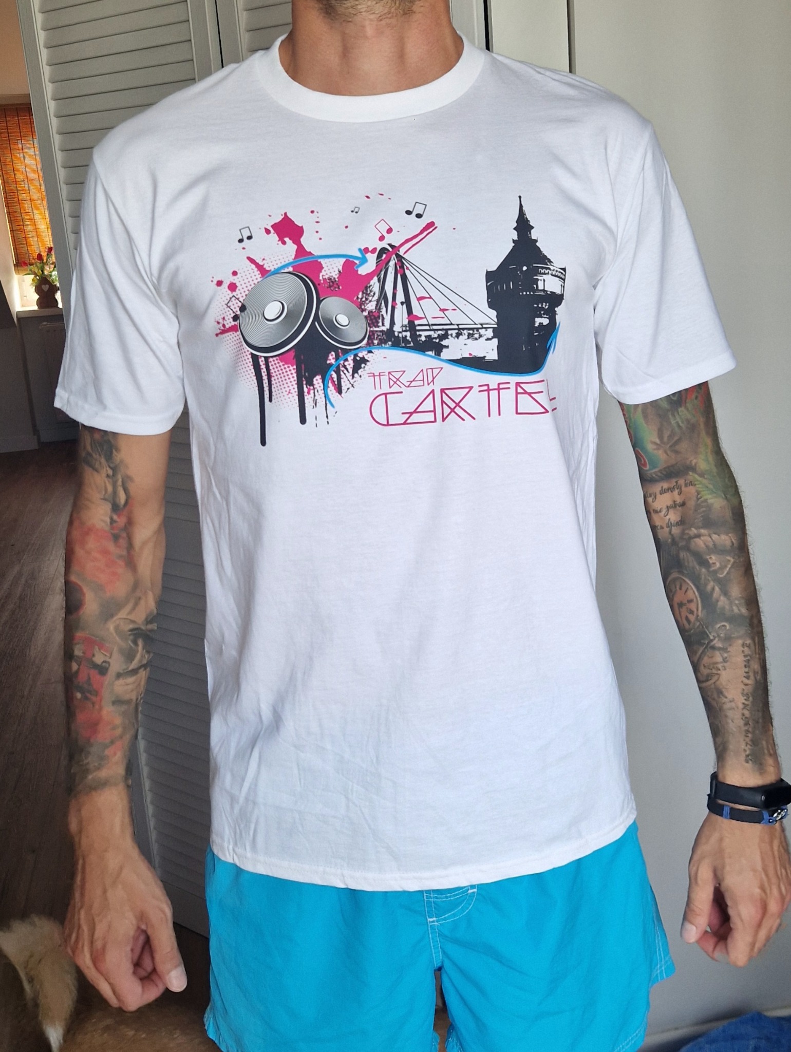 Biała koszulka z unikalnym nadrukiem: wieża, głośniki, nuty i napis 'Trap Cartel'. Mężczyzna w niebieskich szortach prezentuje koszulkę. Widoczne tatuaże na ramionach.