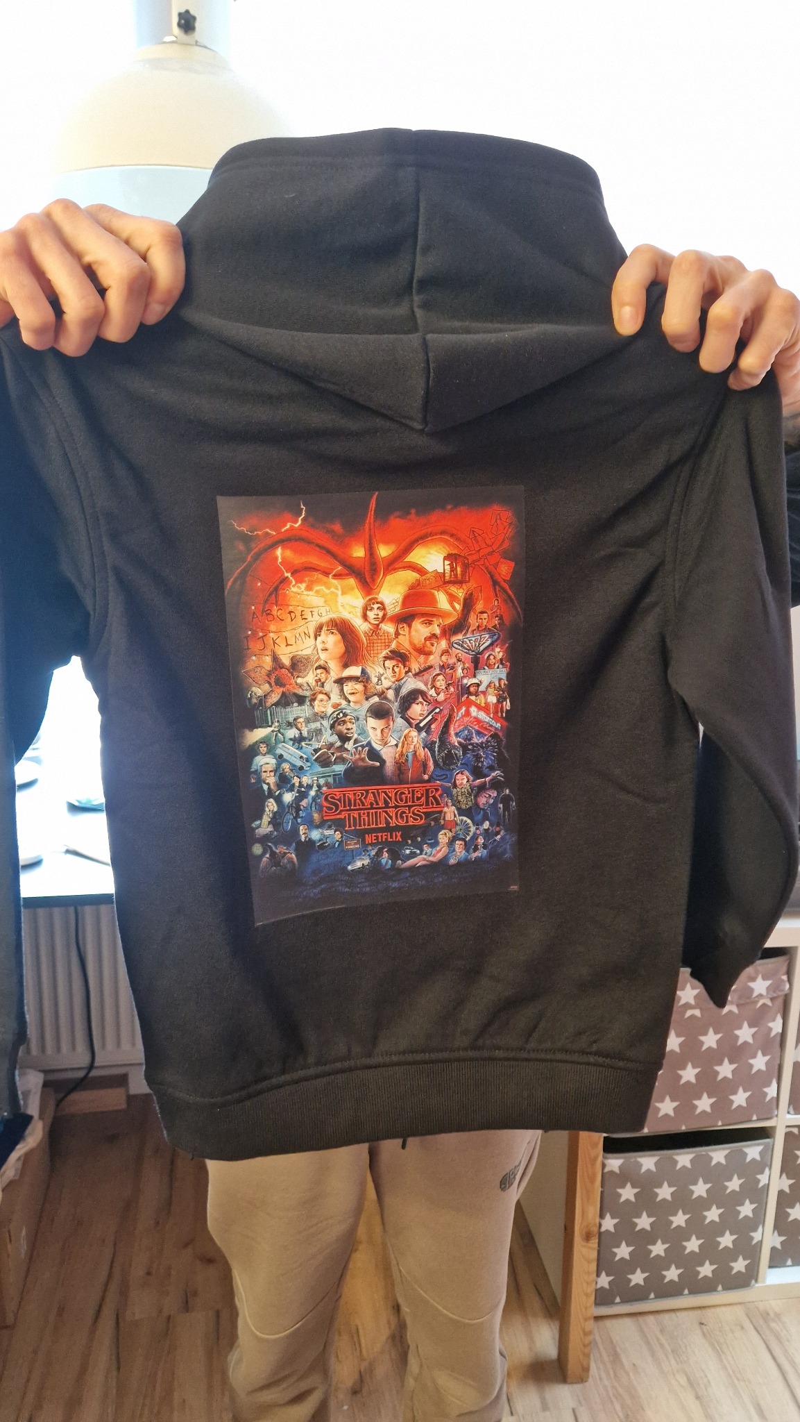 Czarna bluza z kapturem, na plecach nadruk z motywem 'Stranger Things', trzymana przez osobę. Widoczne spodnie dresowe i fragment regału z pudełkami w gwiazdki.