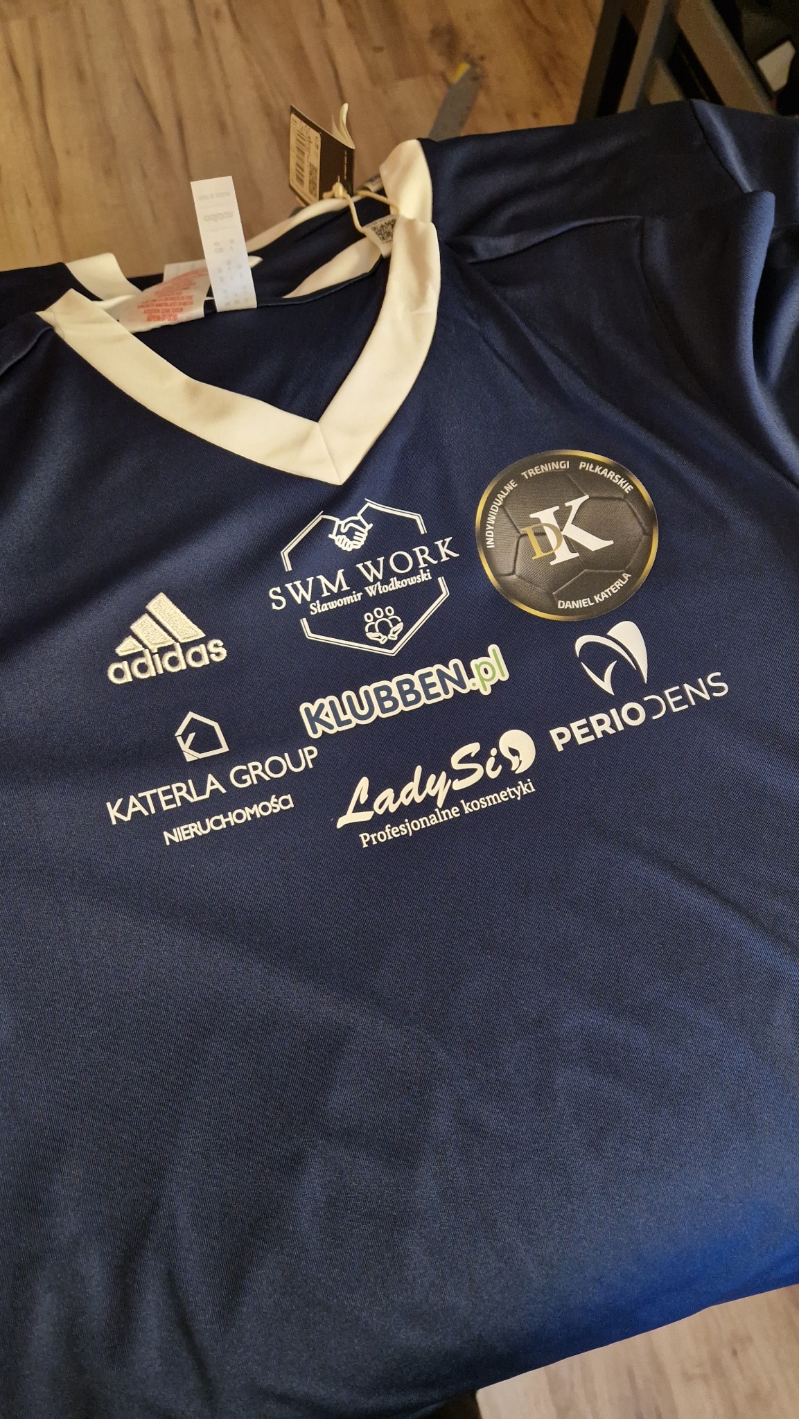 Granatowa koszulka sportowa z białymi nadrukami logo firm, w tym Adidas, Klubben.pl i LadySi. Widoczne metki. Detaliczny widok na nadruki.