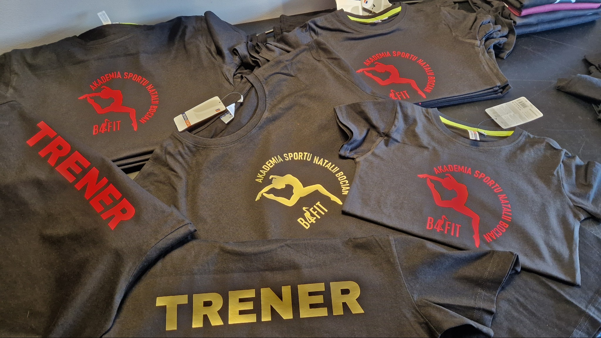 Stos szarych koszulek z nadrukami: logo 'B4FIT' i napisy 'Akademia Sportu Natalii Bocian' oraz 'Trener' w kolorach czerwonym i złotym. Odzież sportowa.