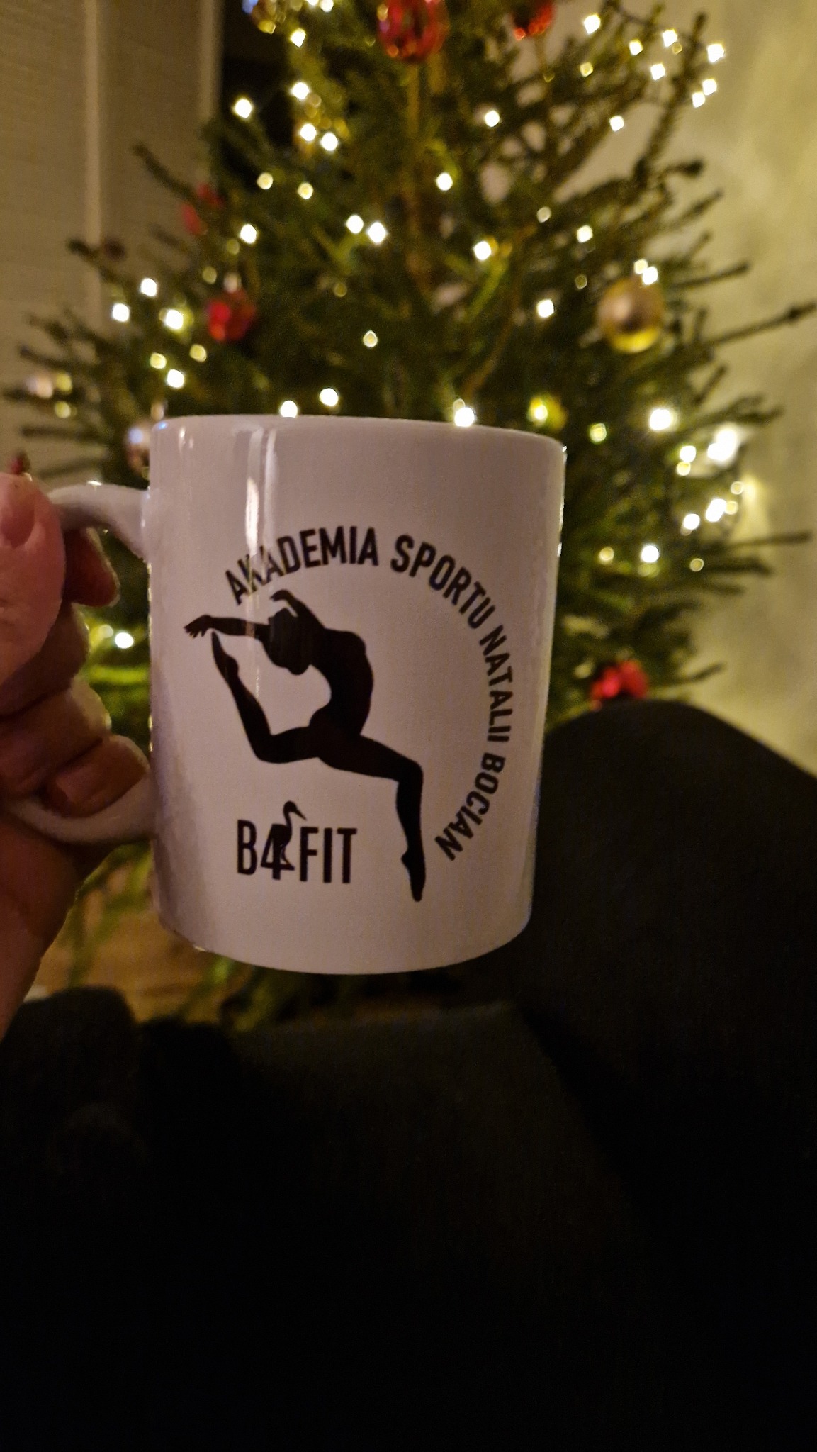 Biały kubek z czarnym nadrukiem logo 'Akademia Sportu Natalii Bocian B4FIT' na tle choinki z lampkami, trzymany w dłoni. Prezentacja identyfikacji wizualnej.