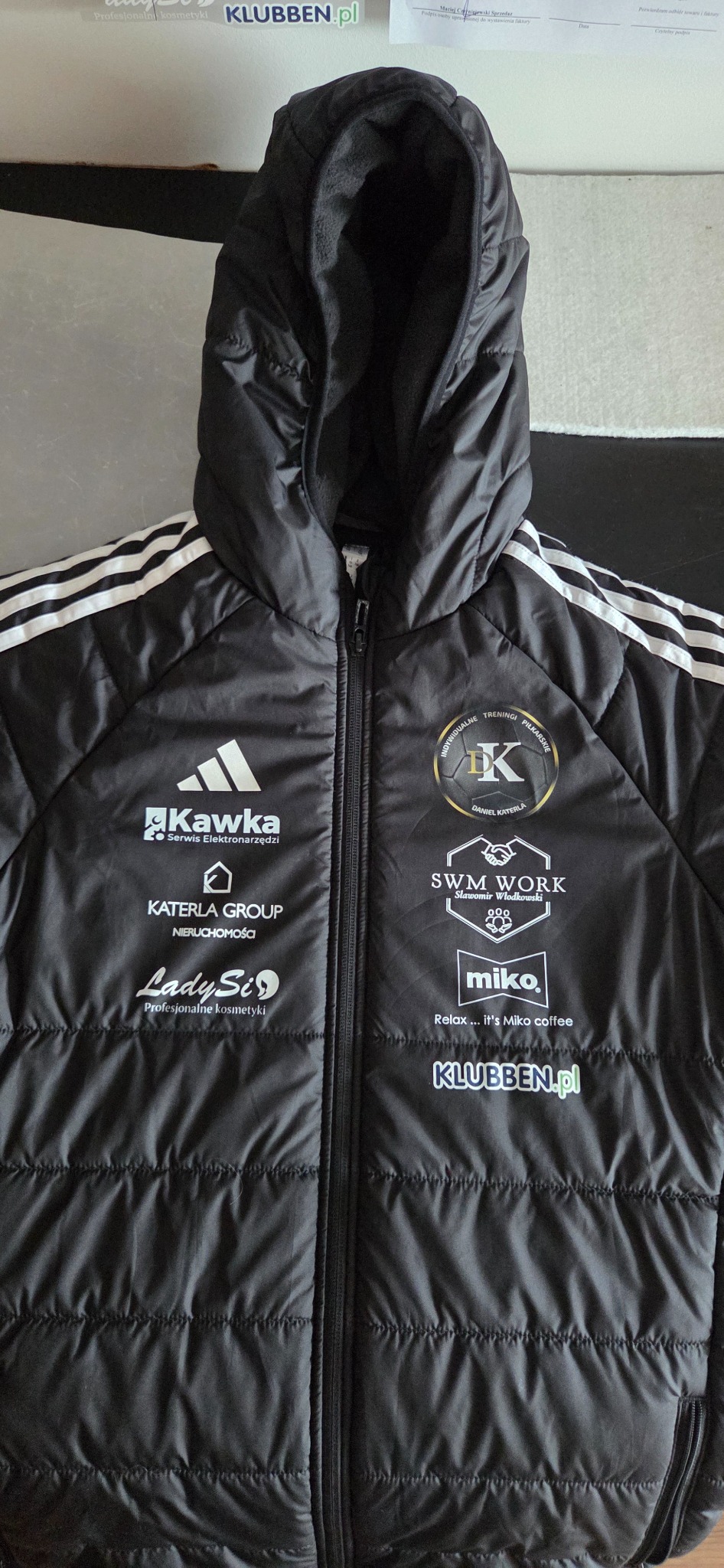 Czarna kurtka puchowa Adidas z nadrukami logo firm i organizacji, w tym Klubben.pl, Kawka Serwis Elektronarzędzi i Katerla Group Nieruchomości.