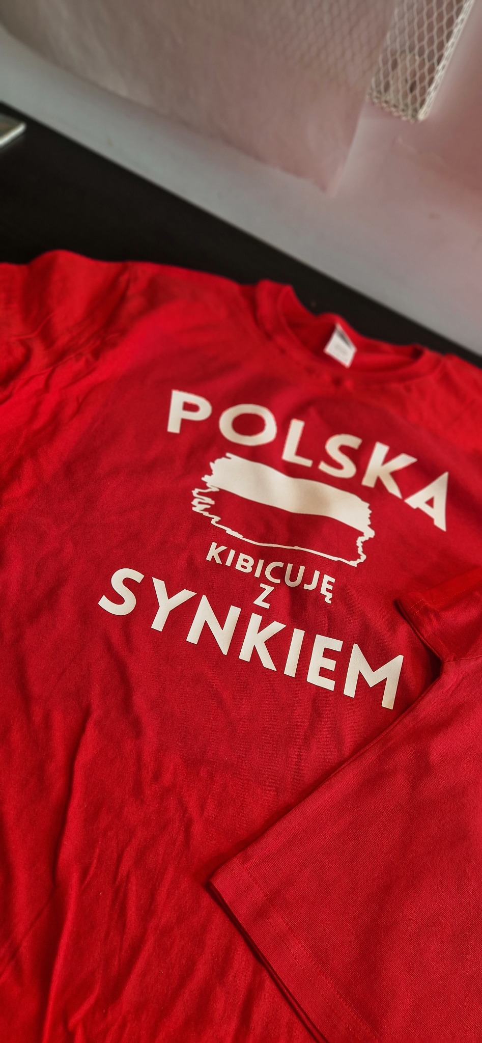 Czerwona koszulka z białym nadrukiem 'Polska Kibicuję z Synkiem', stylizowanym na mapę Polski. Ujęcie z góry, widoczny fragment rękawa.