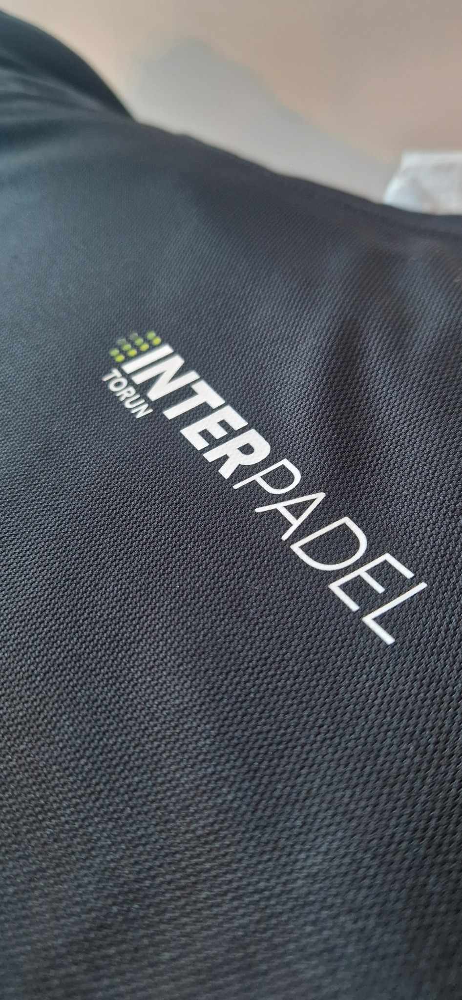 Czarna torba z tkaniny z białym nadrukiem logo 'INTERPADEL TORUŃ' w ukośnym ujęciu, ukazującym strukturę materiału i detale graficzne.