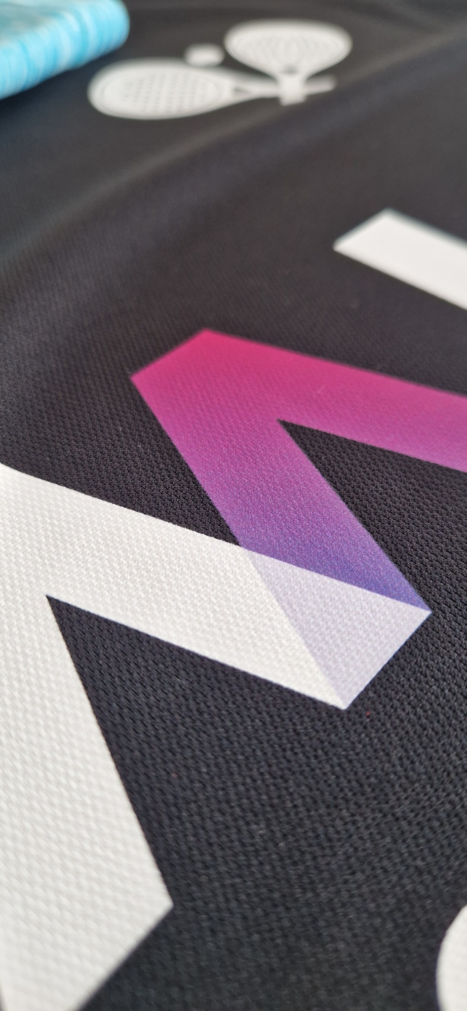 Czarna tkanina z nadrukiem logo w odcieniach bieli i fioletu, zbliżenie na strukturę materiału i detale grafiki. Widoczny fragment uchwytu rakiety tenisowej.