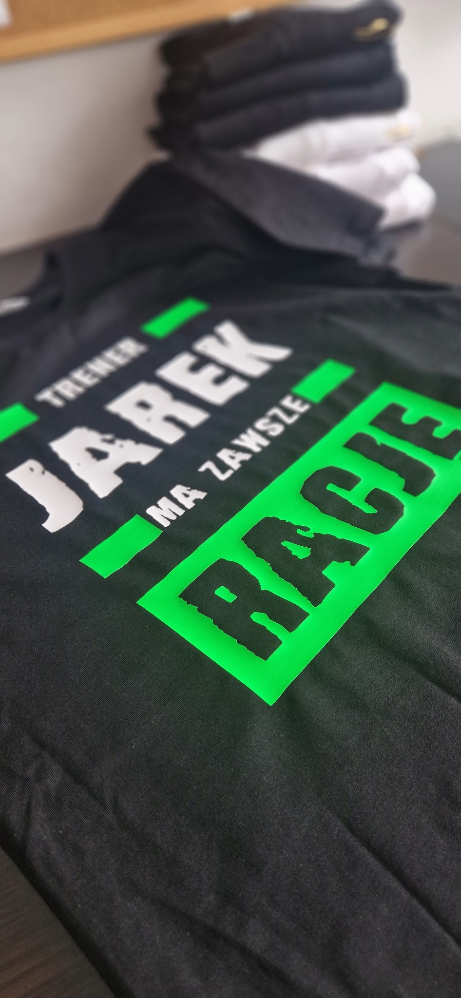 Czarna koszulka z zielonym nadrukiem 'TRENER JAREK MA ZAWSZE RACJE', leżąca na stole, obok stosu innych koszulek. Ujęcie z bliska, pod kątem.