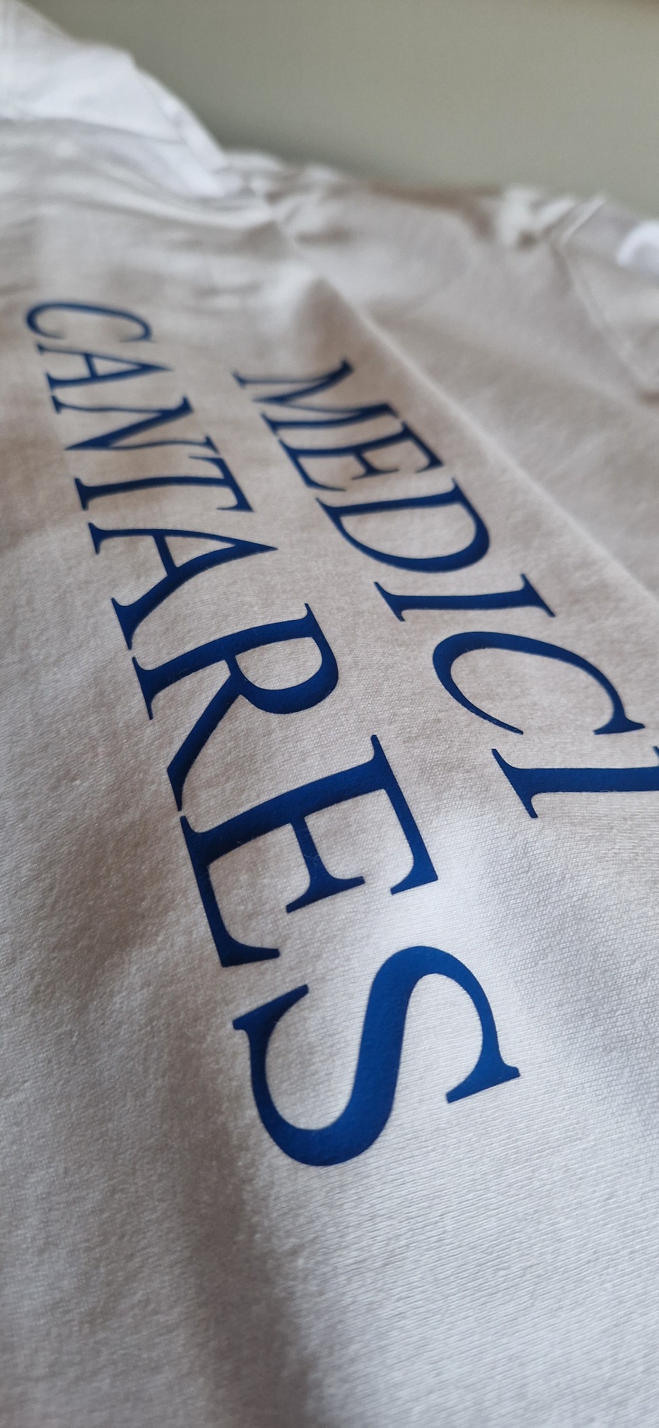 Biała koszulka z niebieskim nadrukiem tekstu 'MEDICI CANTARES' pod kątem, ukazująca detale materiału i precyzję wykonania nadruku.