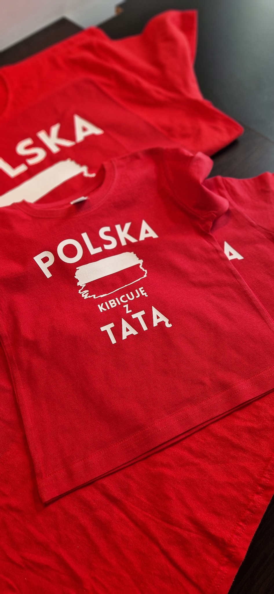 Czerwone koszulki z białym nadrukiem 'Polska Kibicuje z Tatą', leżące na ciemnym blacie. Widoczne trzy sztuki w różnym rozmiarze, ułożone warstwowo.