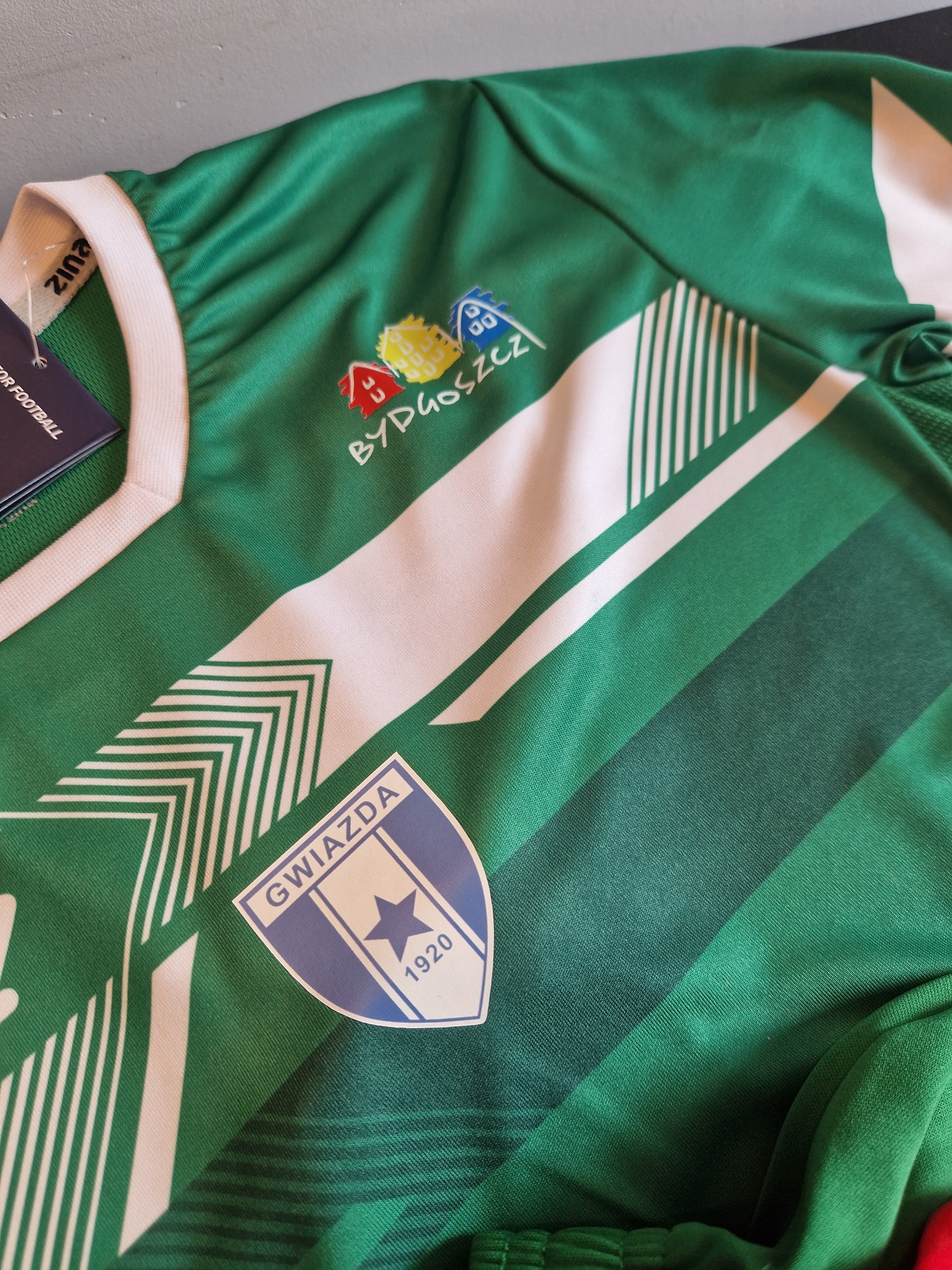 Zielona koszulka sportowa z białymi paskami, herbem 'Gwiazda 1920' i kolorowym logo 'Bydgoszcz' na piersi. Widoczny fragment metki.