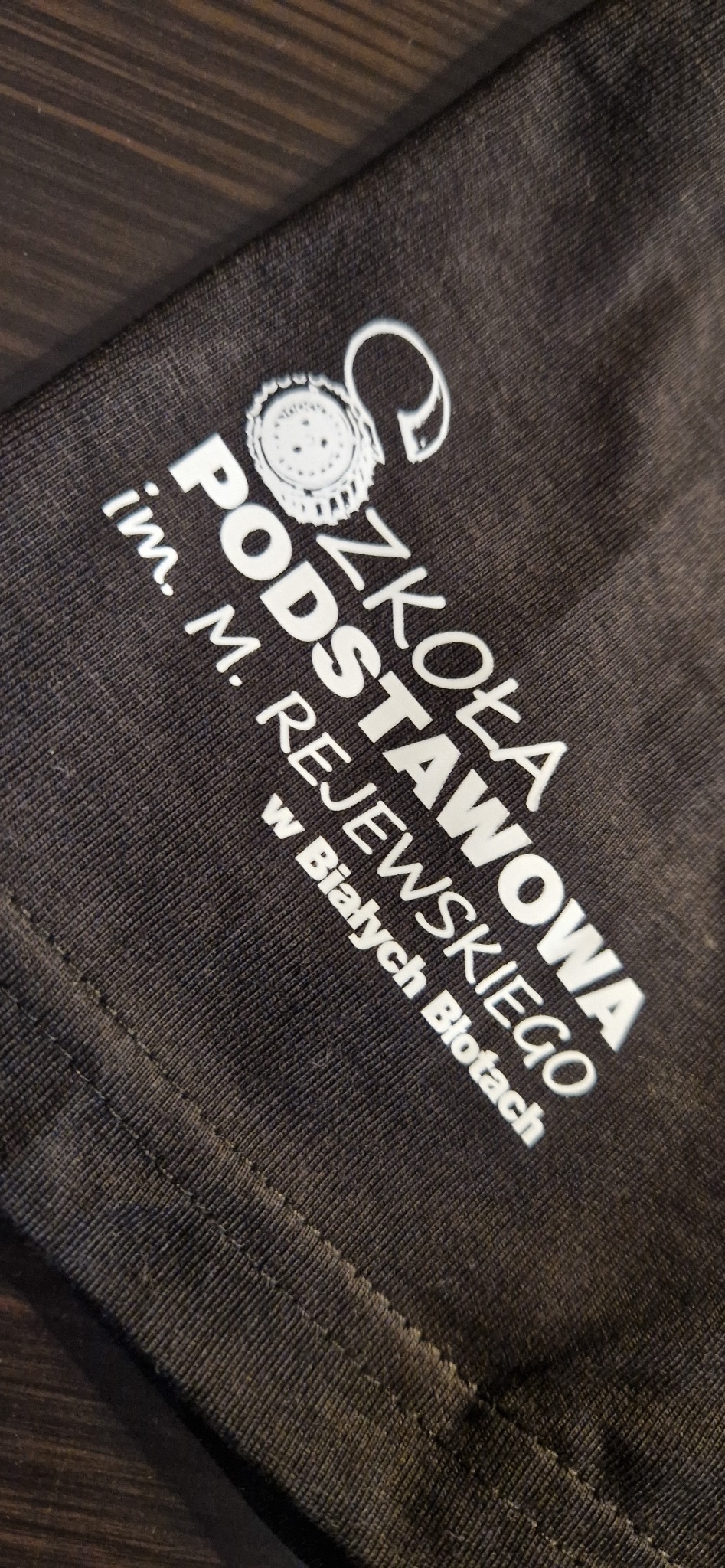 Ciemna koszulka z białym nadrukiem nazwy szkoły i logo, leżąca na drewnianym stole. Widoczny fragment szwu koszulki.