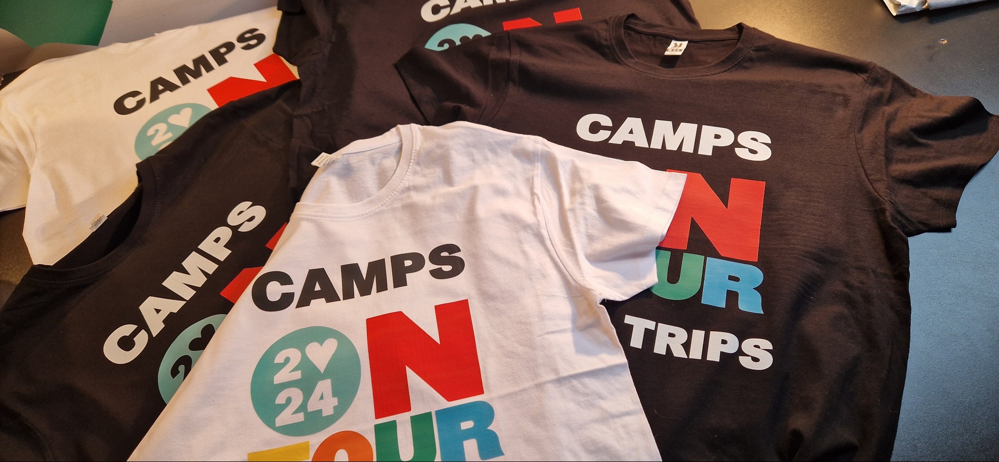 Stos koszulek z nadrukiem 'Camps on tour 2024' w kolorach białym i czarnym, ułożonych na ciemnym tle. Nadruk w stylu graficznym, z użyciem różnych kolorów.