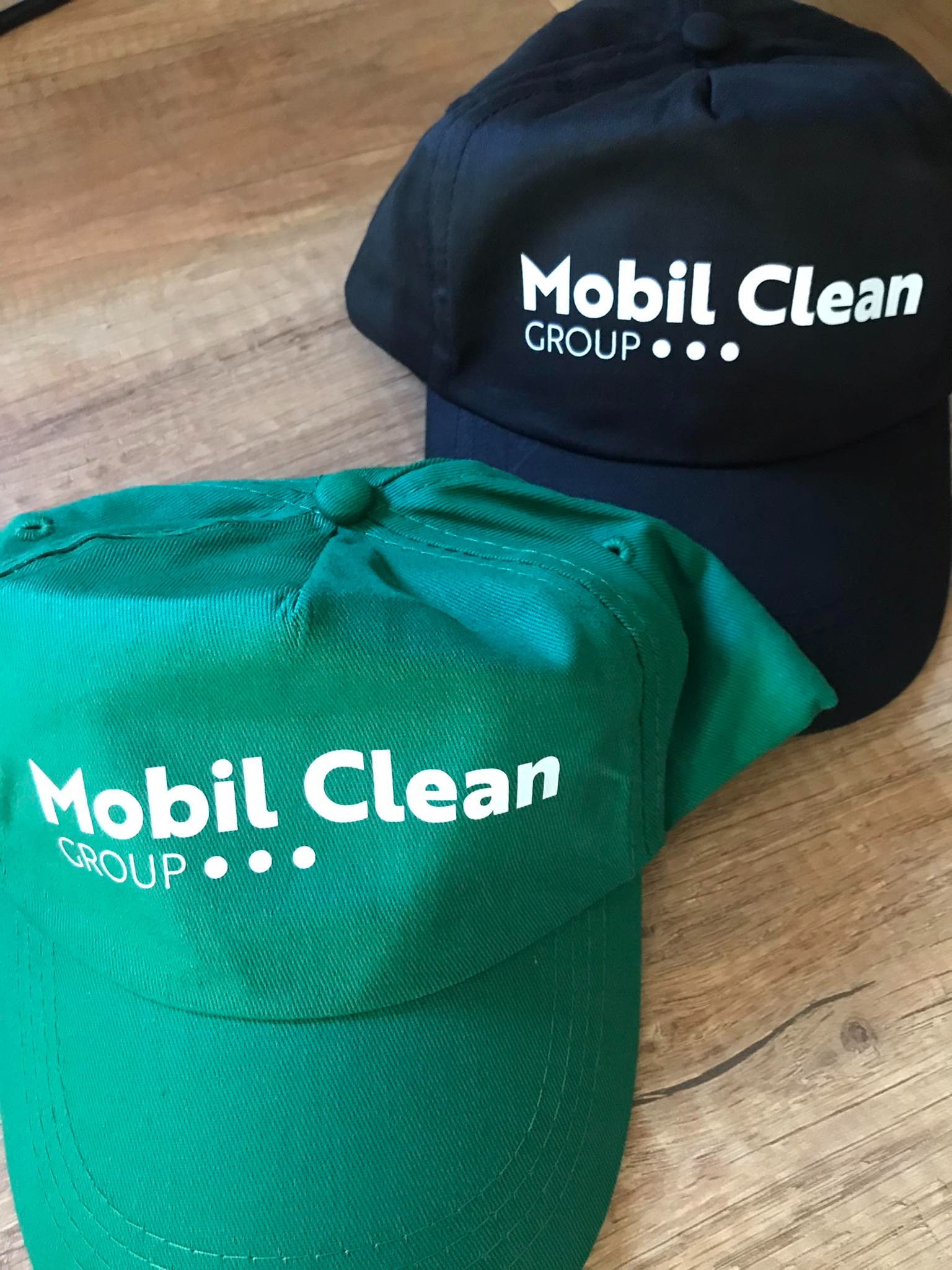 Dwie czapki z daszkiem: zielona i czarna, z białym nadrukiem 'Mobil Clean GROUP', leżące na drewnianej powierzchni. Widoczne detale szwów i faktura materiału.