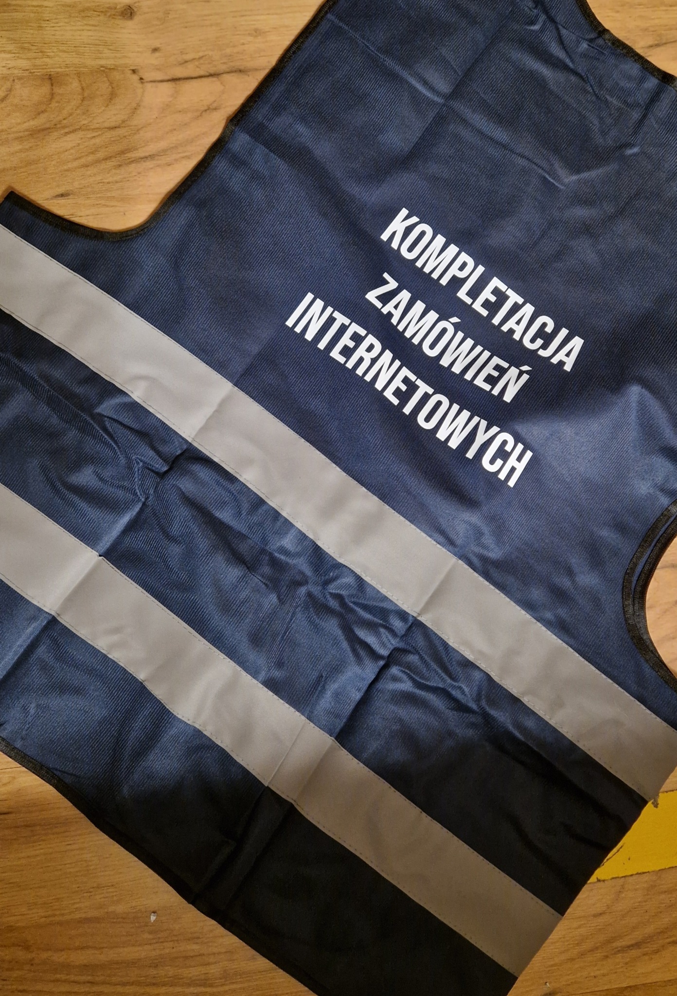 Granatowa kamizelka odblaskowa z białym napisem 'Kompletacja zamówień internetowych' leżąca na drewnianym podłożu, widok z góry.