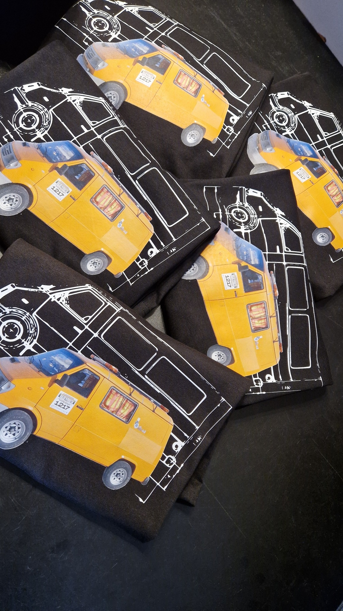 Stos czarnych koszulek z nadrukiem żółtego samochodu na tle białego schematu, ułożonych pod kątem na ciemnej powierzchni. Nadruk wykonany metodą sitodruku.