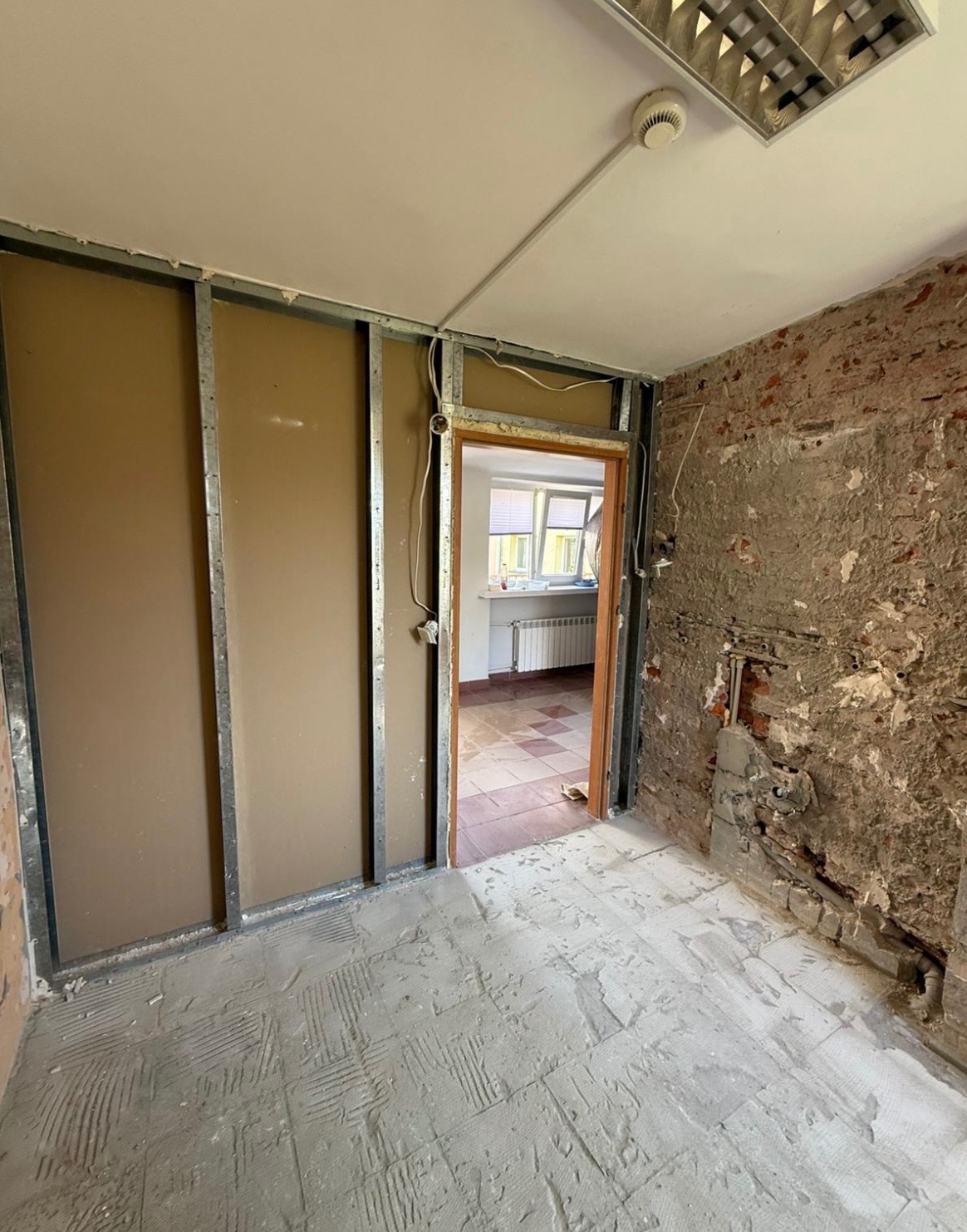 Wnętrze w trakcie remontu: odsłonięta cegła, metalowe profile ścian działowych, surowa podłoga i widok na sąsiednie pomieszczenie z grzejnikiem.