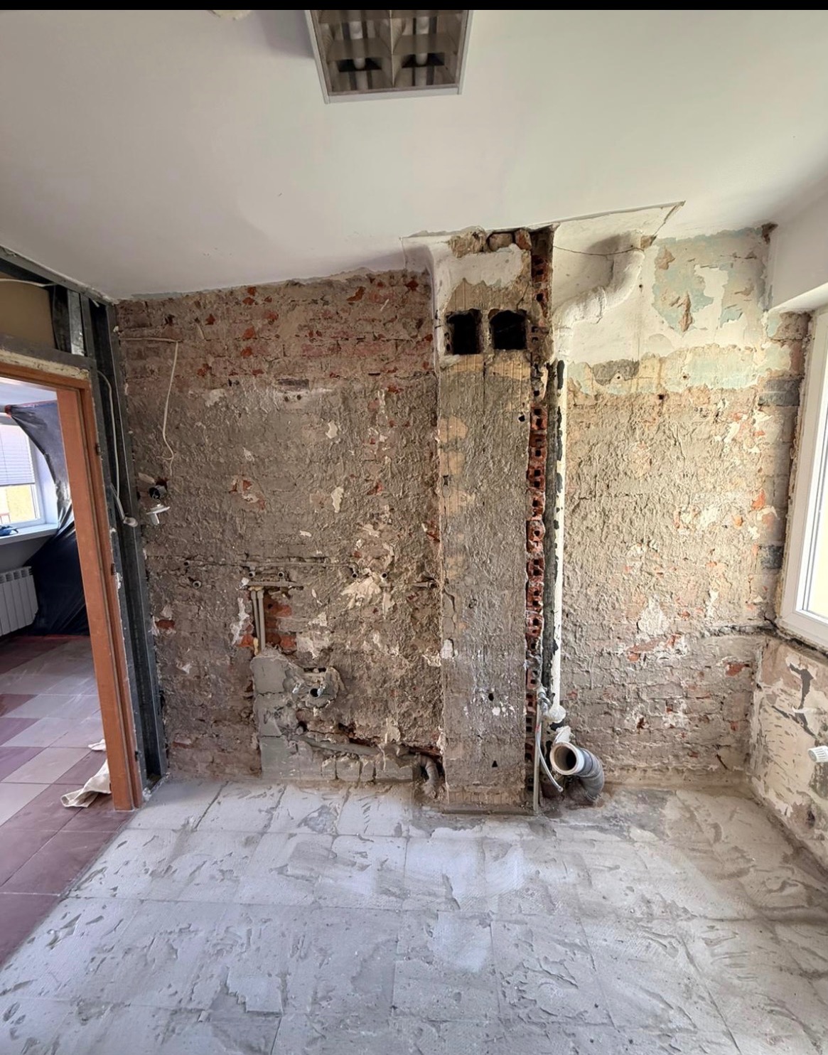 Odsłonięta cegła i instalacje po wyburzeniu ściany w pomieszczeniu z częściowo usuniętą podłogą. Widoczne rury i otwory wentylacyjne.
