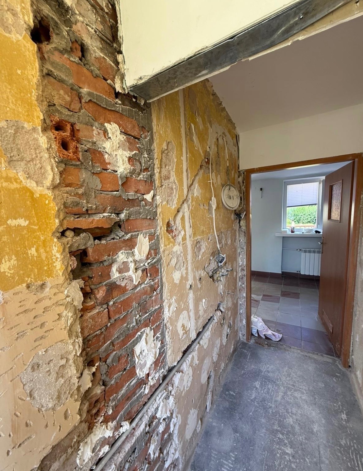 Odsłonięta cegła i resztki tapety po wyburzeniu ściany w korytarzu. Widoczne przewody elektryczne i fragment futryny drzwi. Podłoga betonowa, w tle widok na pokój.