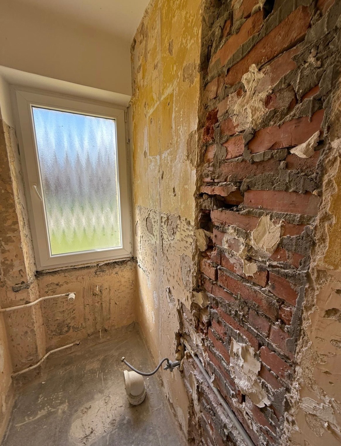 Częściowo wyburzona ściana z czerwonej cegły w pomieszczeniu z oknem. Widoczne instalacje i resztki tynku na ścianach. Etap prac wyburzeniowych.