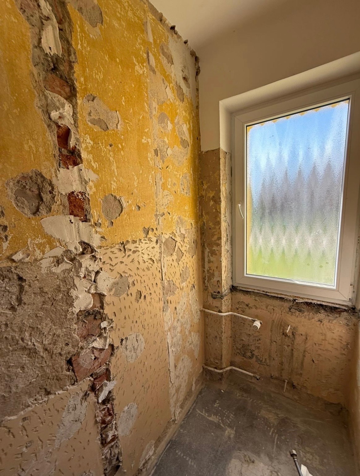 Wnętrze łazienki w trakcie remontu: ściany z resztkami starej farby i tynku, odsłonięta cegła, okno z widokiem na zewnątrz. Widoczne rury instalacji wodnej.