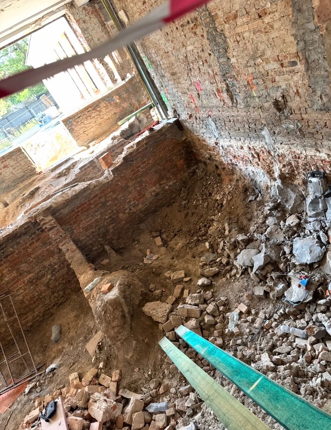 Wnętrze budynku w trakcie rozbiórki, widoczne fragmenty ceglanych ścian, gruz i drewniane elementy. Ujęcie z góry, z widoczną taśmą ostrzegawczą.