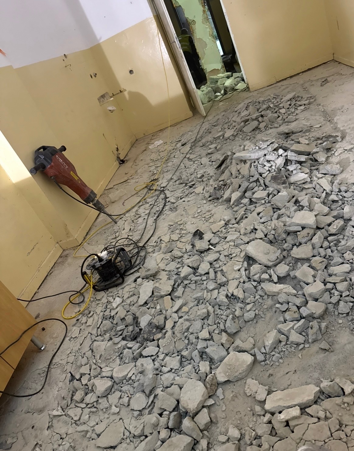 Pokój po wyburzeniu, gruz na podłodze, widoczny młot pneumatyczny przy ścianie. Przejście do kolejnego pomieszczenia z resztkami gruzu.