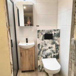 BOGUSŁAW KROPACZ - Wykończona łazienka w Krakowie z kabiną prysznicową, umywalką z szafką i toaletą. Na ścianie płytki z motywem roślinnym i lustro, w którym odbija się osoba.