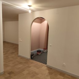 BOGUSŁAW KROPACZ - Wnętrze po malowaniu: jasne ściany, parkiet, widoczne puszki farb Flugger w przejściu z łukiem. Świeżo odmalowane pomieszczenie z widocznymi detalami wykończeniowymi.