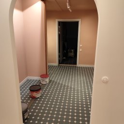 BOGUSŁAW KROPACZ - Wnętrze po malowaniu z łukowym przejściem, widoczne puszki z farbą Flugger, szaro-biała podłoga w romby i różowe ściany. Oświetlenie punktowe.