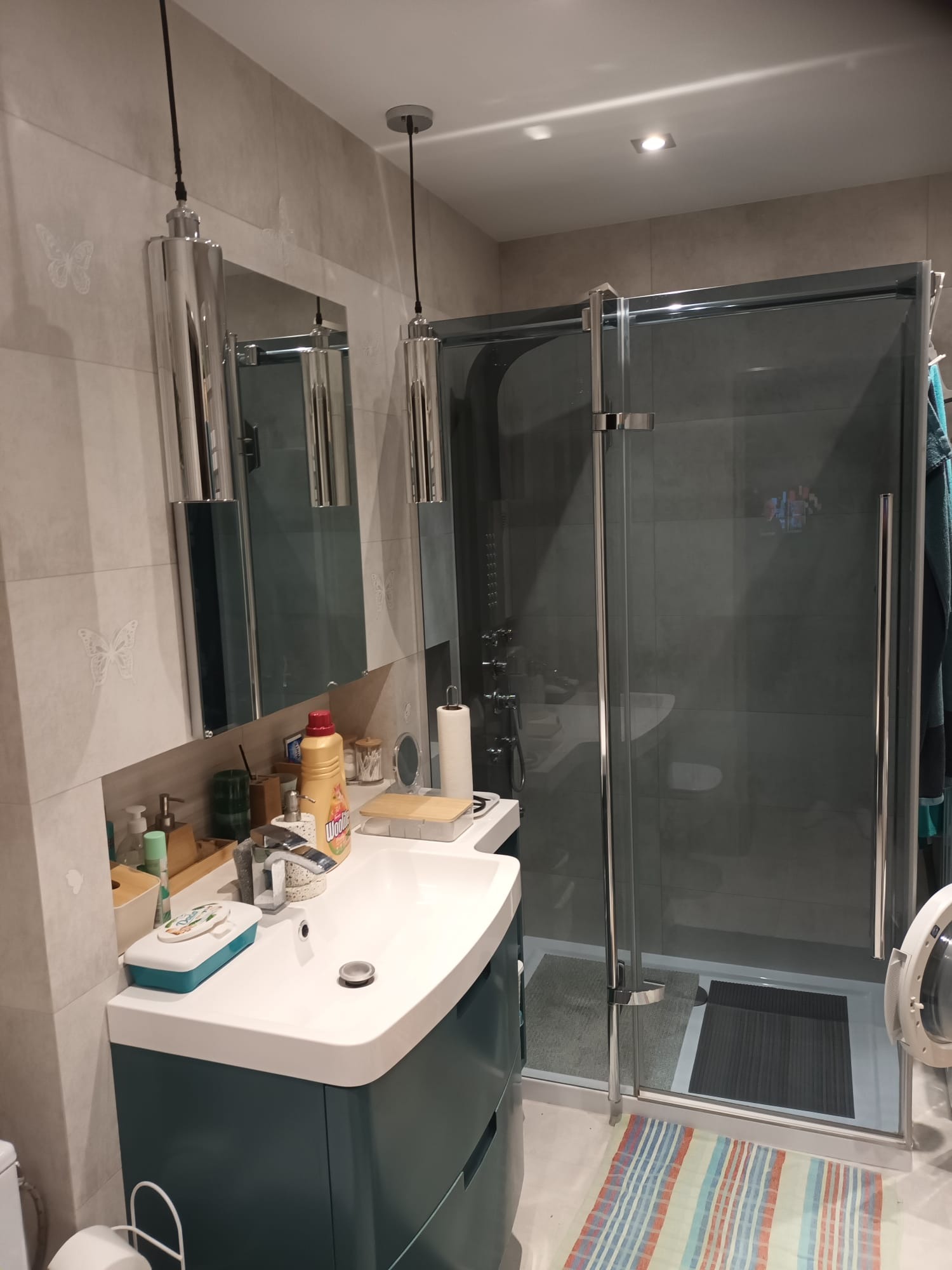 Nowoczesna łazienka po remoncie: kabina prysznicowa ze szklanymi drzwiami, umywalka z szafką w kolorze butelkowej zieleni, lustro i lampy wiszące. Na ścianach płytki z motywem motyli.