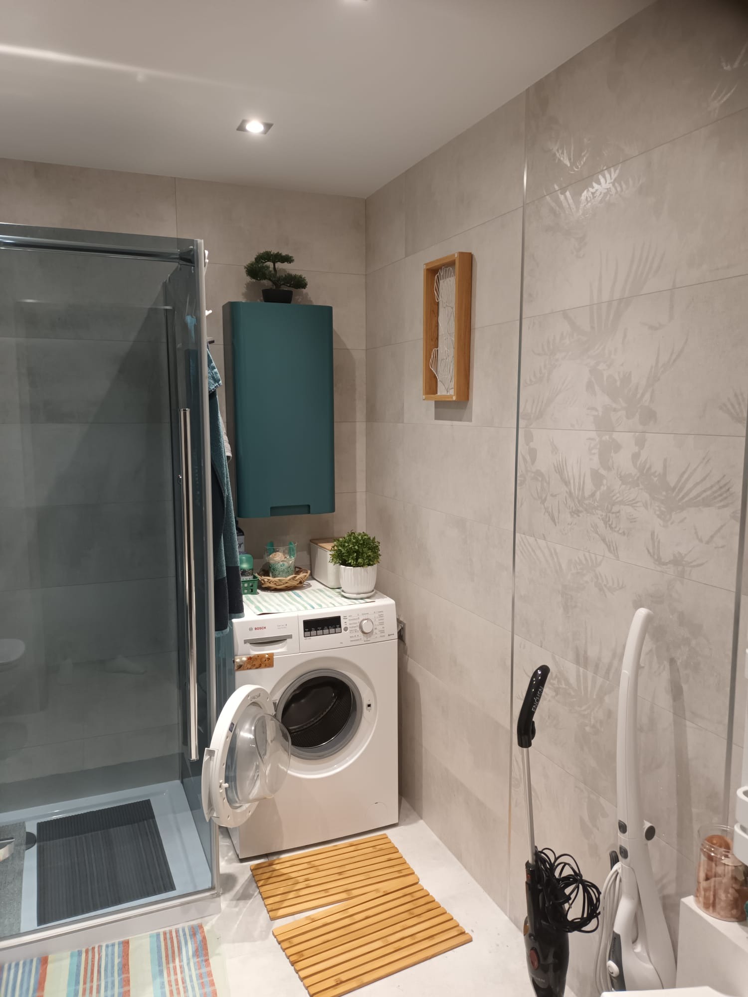 Jasna łazienka po remoncie z kabiną prysznicową, pralką Bosch, szafką w kolorze morskim i płytkami z motywem roślinnym. Widoczne akcesoria i dekoracje.