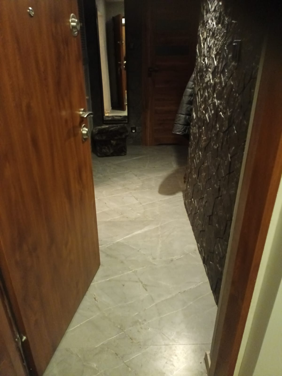 Wykończony korytarz z marmurową podłogą i dekoracyjną ścianą z kamienia. Otwarta drewniana dębowa drzwi wejściowe. W tle widoczne lustro i elementy garderoby.