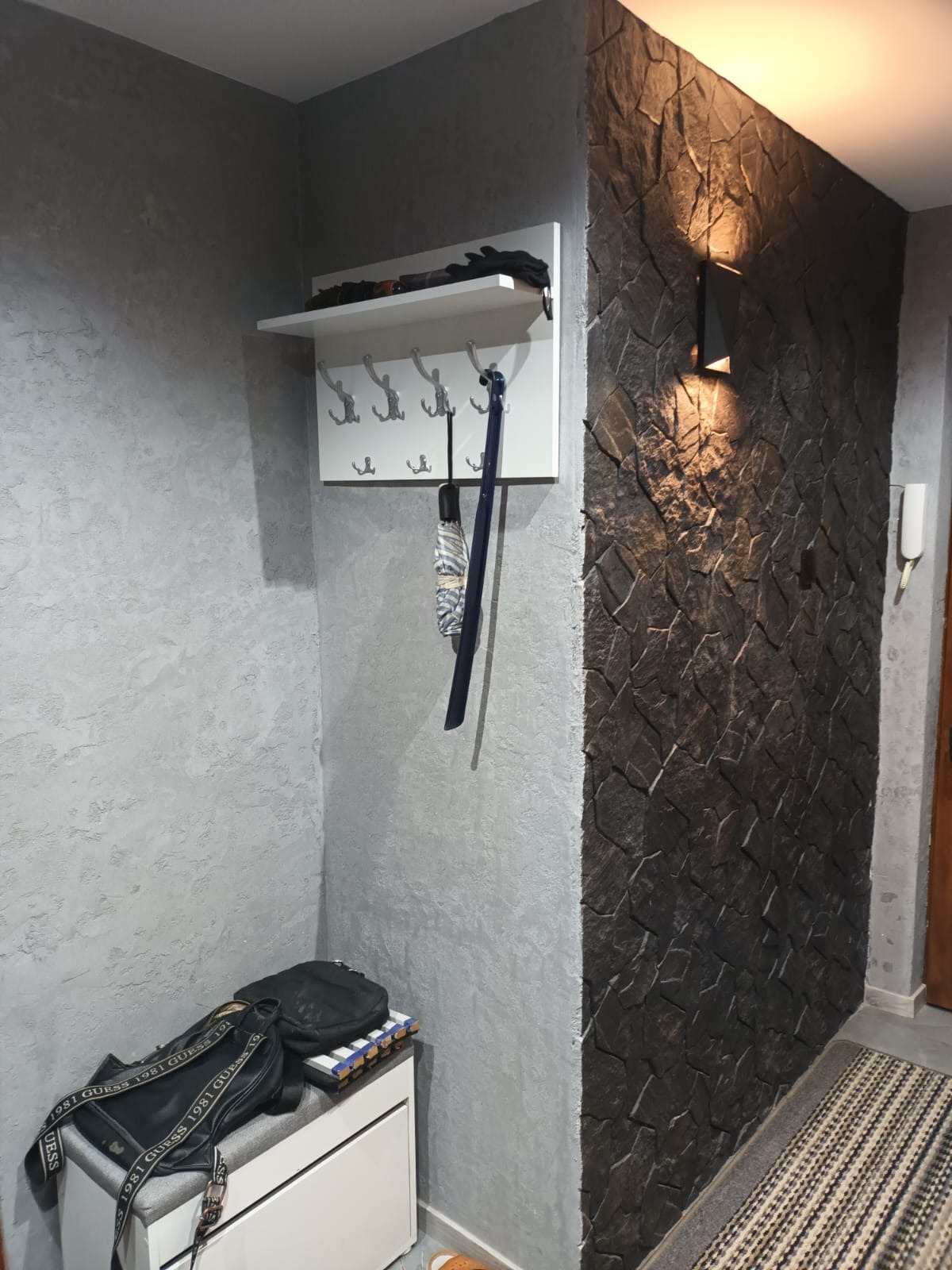 Nowoczesny przedpokój z szarą, teksturowaną ścianą i dekoracyjną ścianą z kamienia. Biały wieszak z półką, torby na pufie. Oświetlenie punktowe.