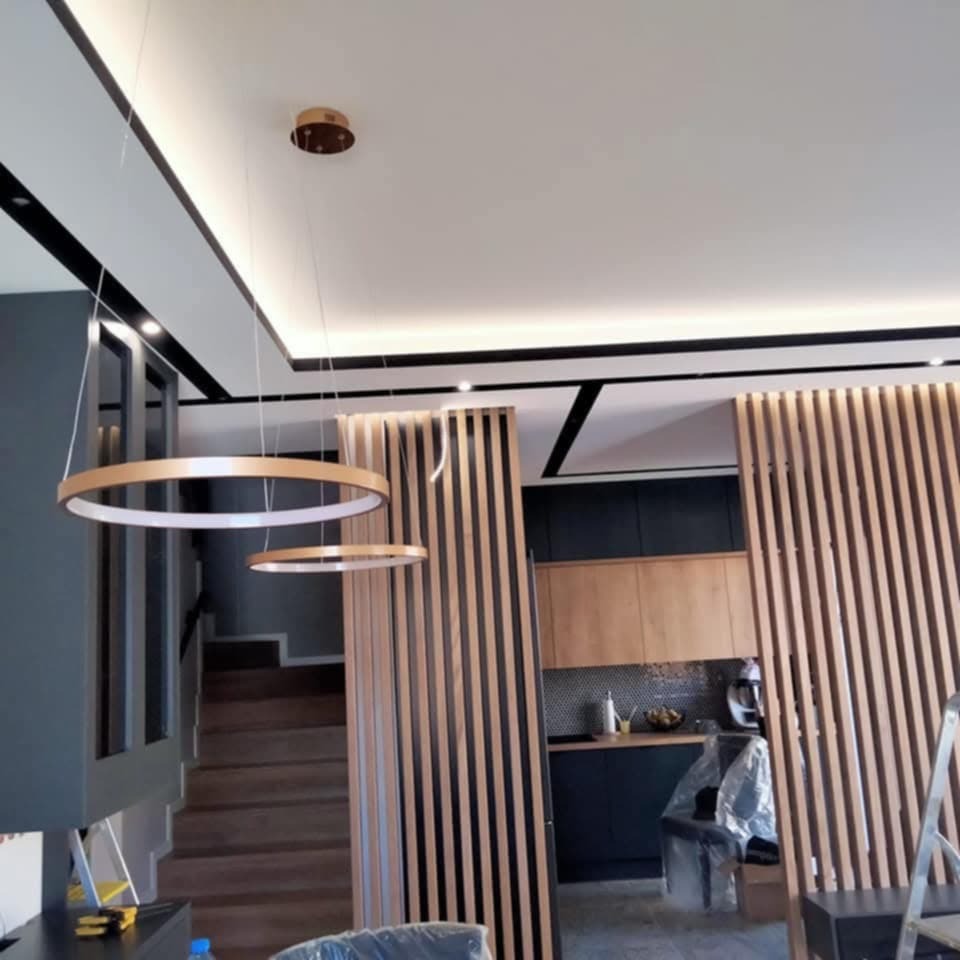 Nowoczesne wnętrze z okrągłymi lampami wiszącymi, drewnianą ścianką działową i schodami w tle. Widoczna kuchnia z drewnianymi szafkami i czarnymi elementami.