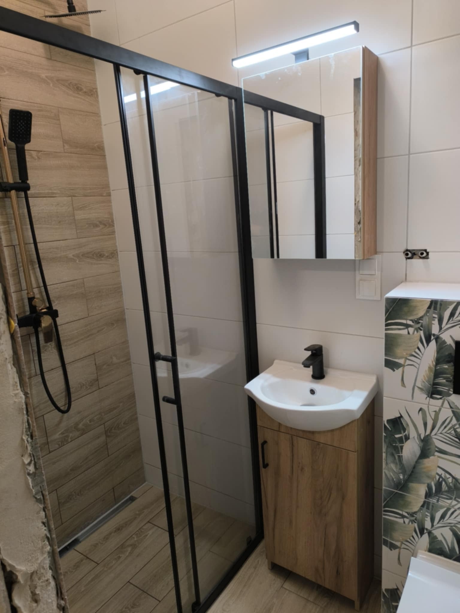 Wykończona łazienka z kabiną prysznicową typu walk-in z czarnymi profilami, umywalką z szafką w kolorze drewna i płytkami z motywem roślinnym.