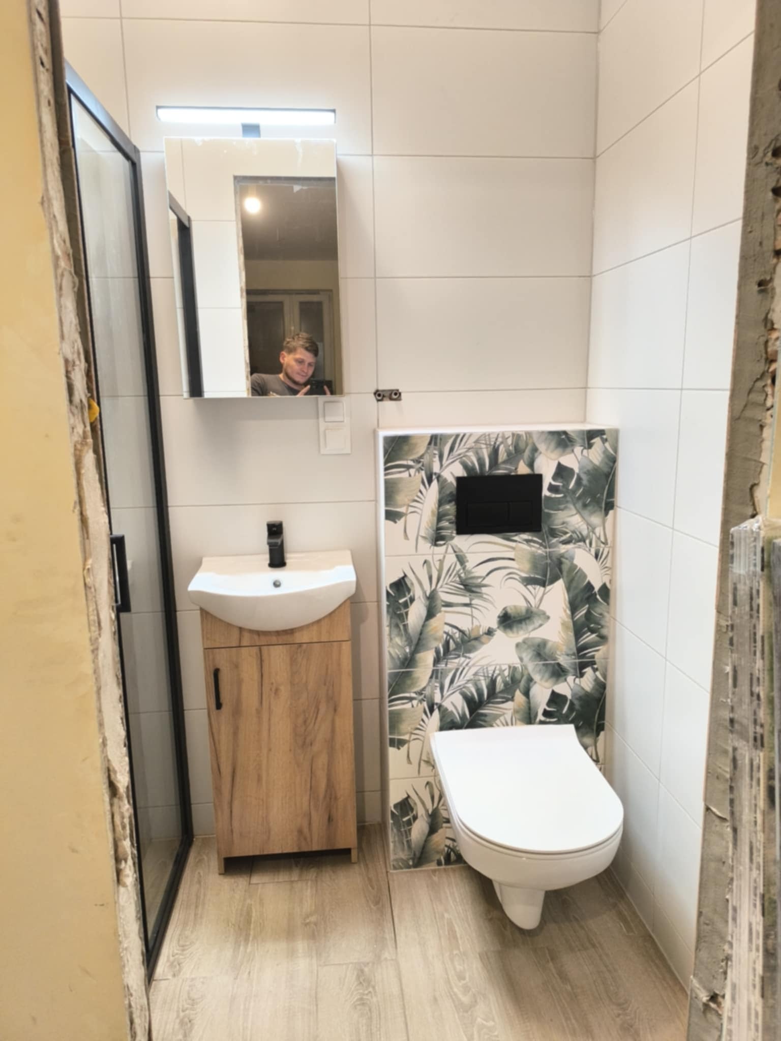 Wykończona łazienka w Krakowie z kabiną prysznicową, umywalką z szafką i toaletą. Na ścianie płytki z motywem roślinnym i lustro, w którym odbija się osoba.