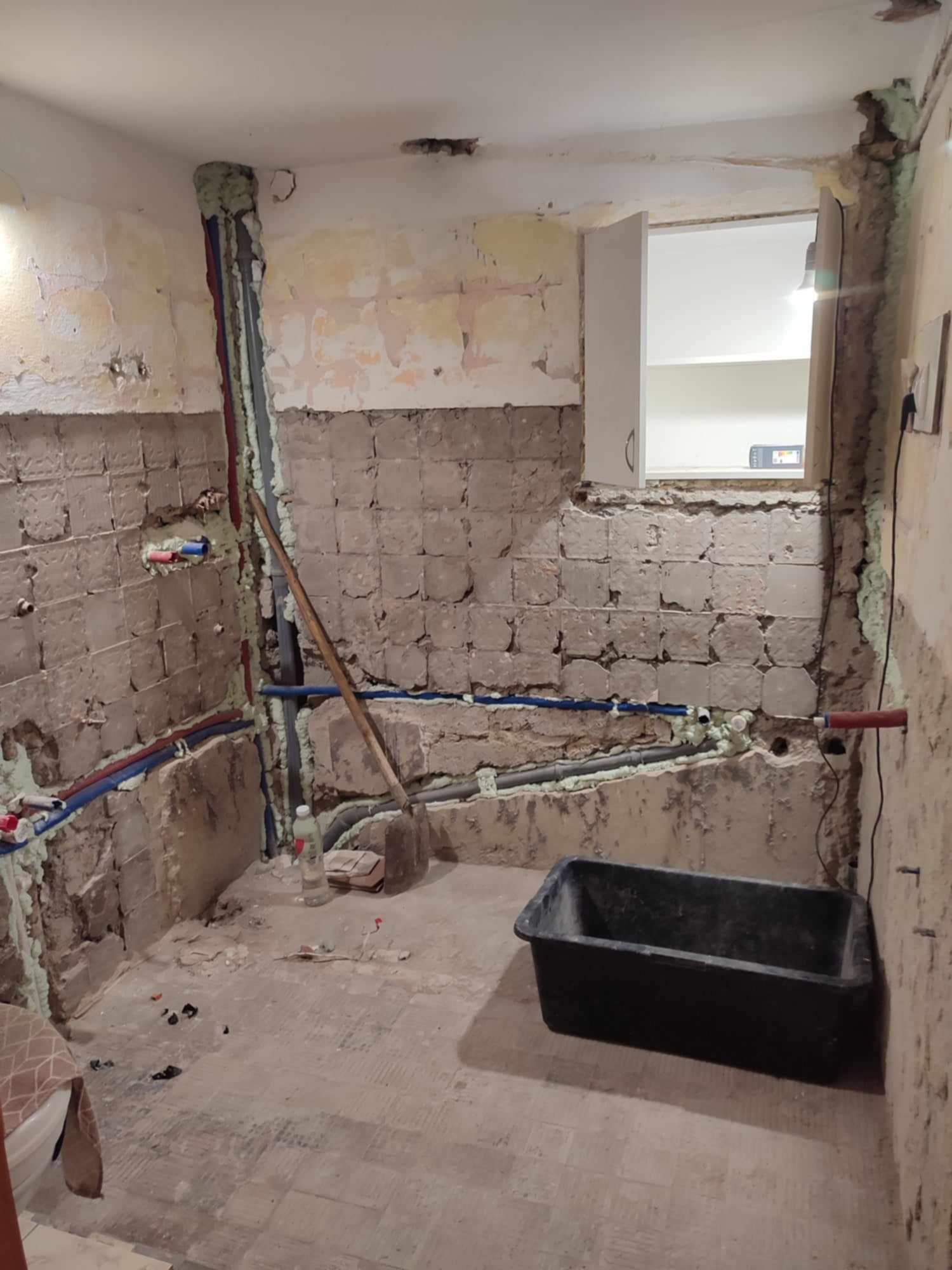 Łazienka w trakcie remontu: częściowo usunięte płytki, widoczne rury zabezpieczone pianką montażową, czarne wiadro na podłodze, okno z otwartą szafką w tle.