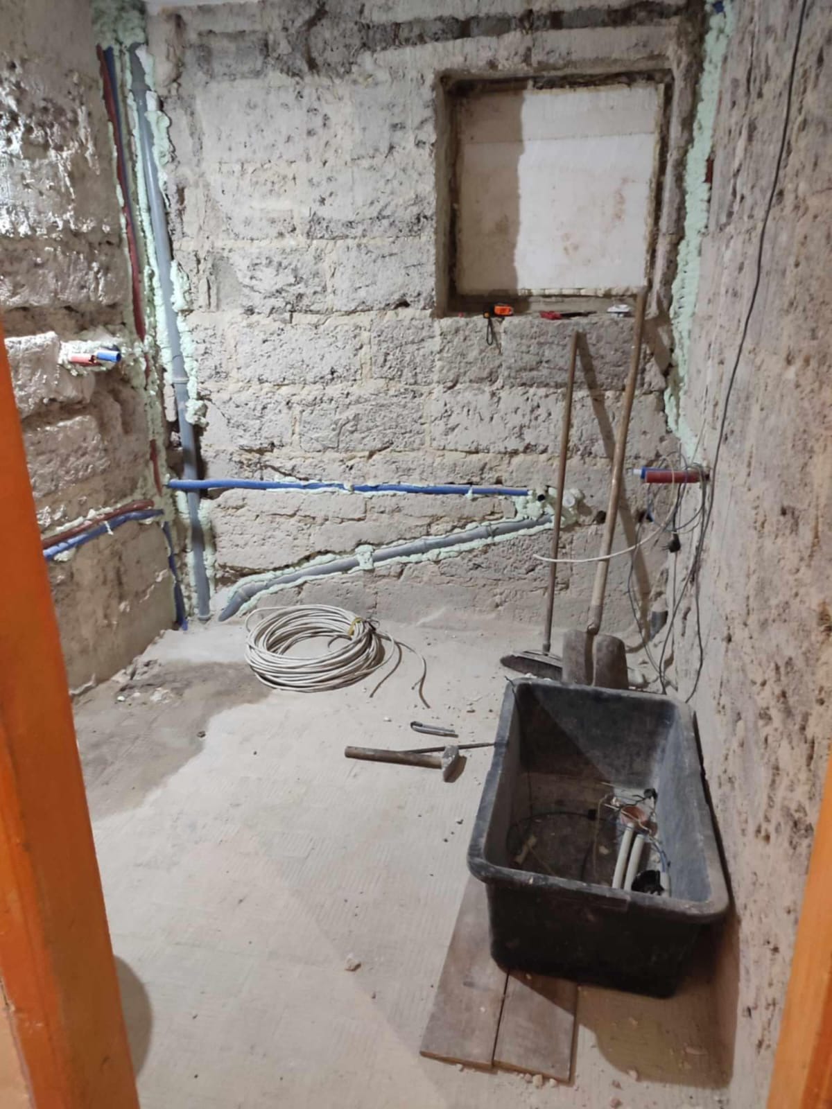 Surowy stan pomieszczenia w trakcie remontu: instalacje wodne zabezpieczone pianką, narzędzia, taczka. Ściany z cegły, okno zamurowane. Widok zza framugi.