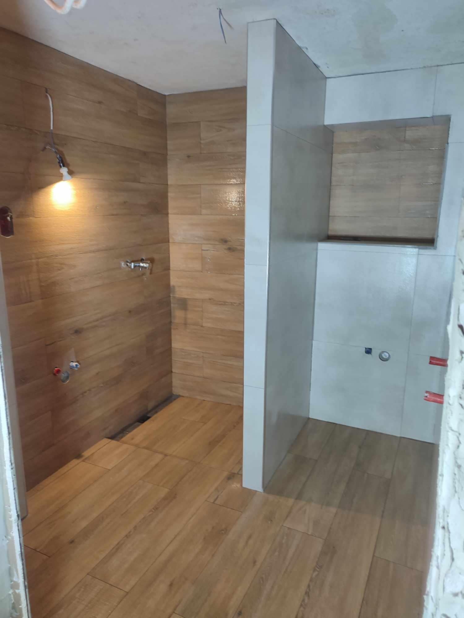 Wykończona kabina prysznicowa z drewnopodobnymi i szarymi płytkami, widoczne instalacje wodne i oświetlenie. Częściowo wykończona łazienka w trakcie remontu.