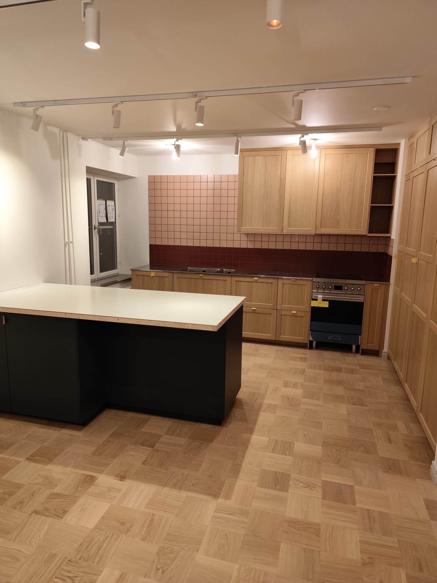 Jasna kuchnia z drewnianymi szafkami, wyspą z ciemnym blatem i parkietem w jodełkę. Ściana nad blatem wyłożona różowo-bordową mozaiką. Oświetlenie szynowe.