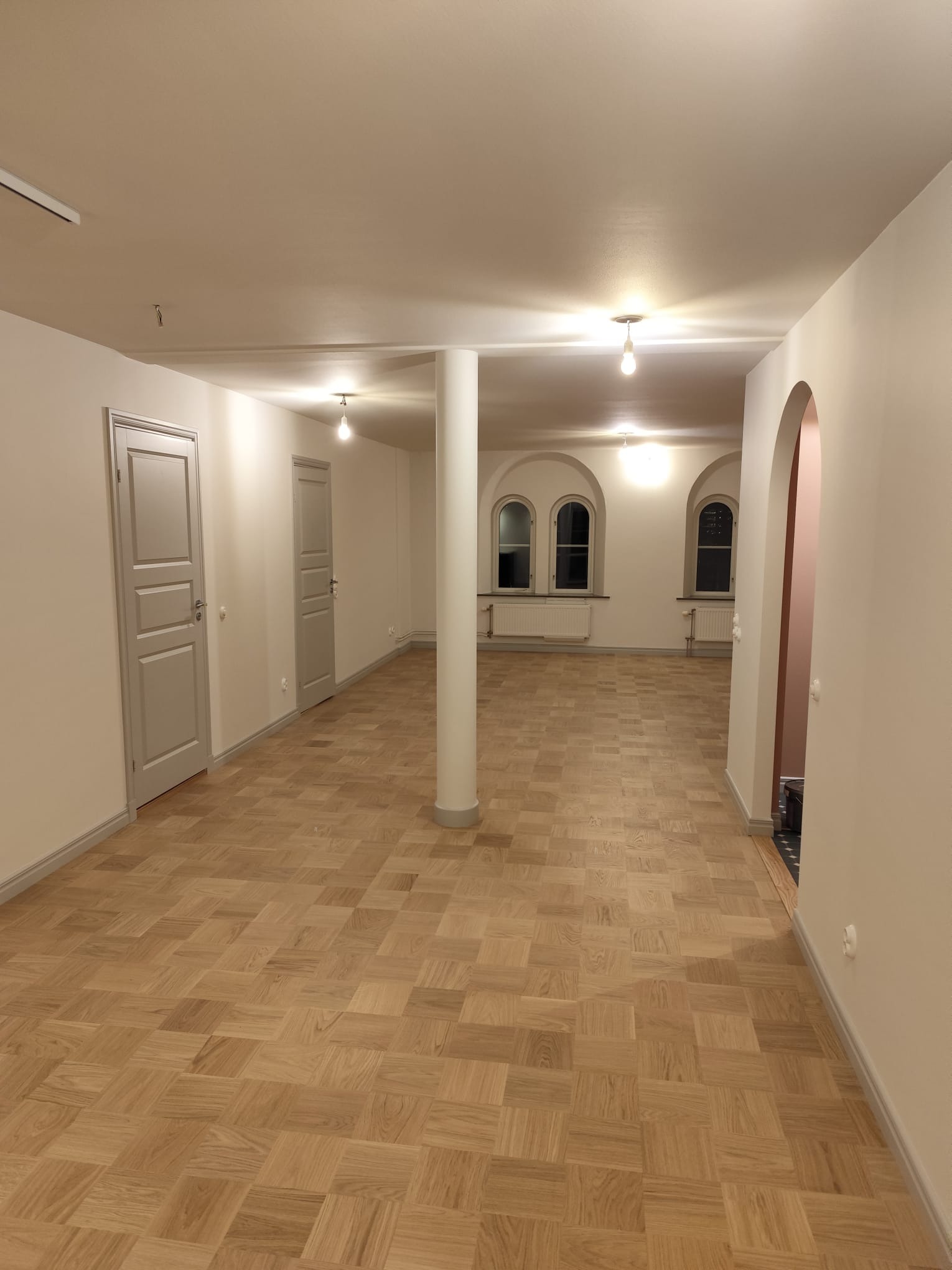 Jasne, przestronne wnętrze po remoncie z parkietem w jodełkę, kolumną i szarymi drzwiami. Widoczne okna łukowe i świeżo pomalowane ściany.