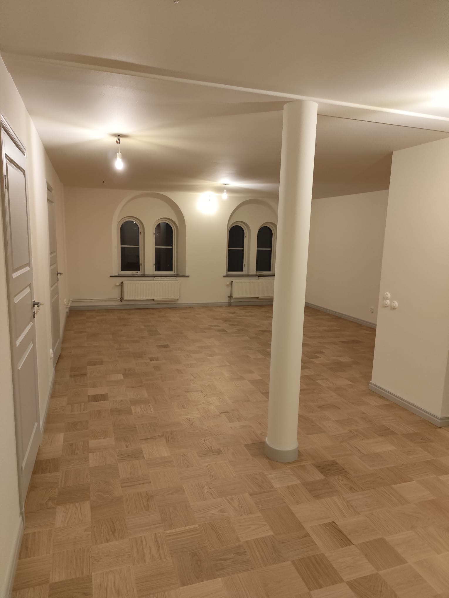 Jasny parkiet w jodełkę francuską w przestronnym, odnowionym wnętrzu z kolumną i oknami łukowymi. Świeżo pomalowane ściany i widoczne gniazdka elektryczne.
