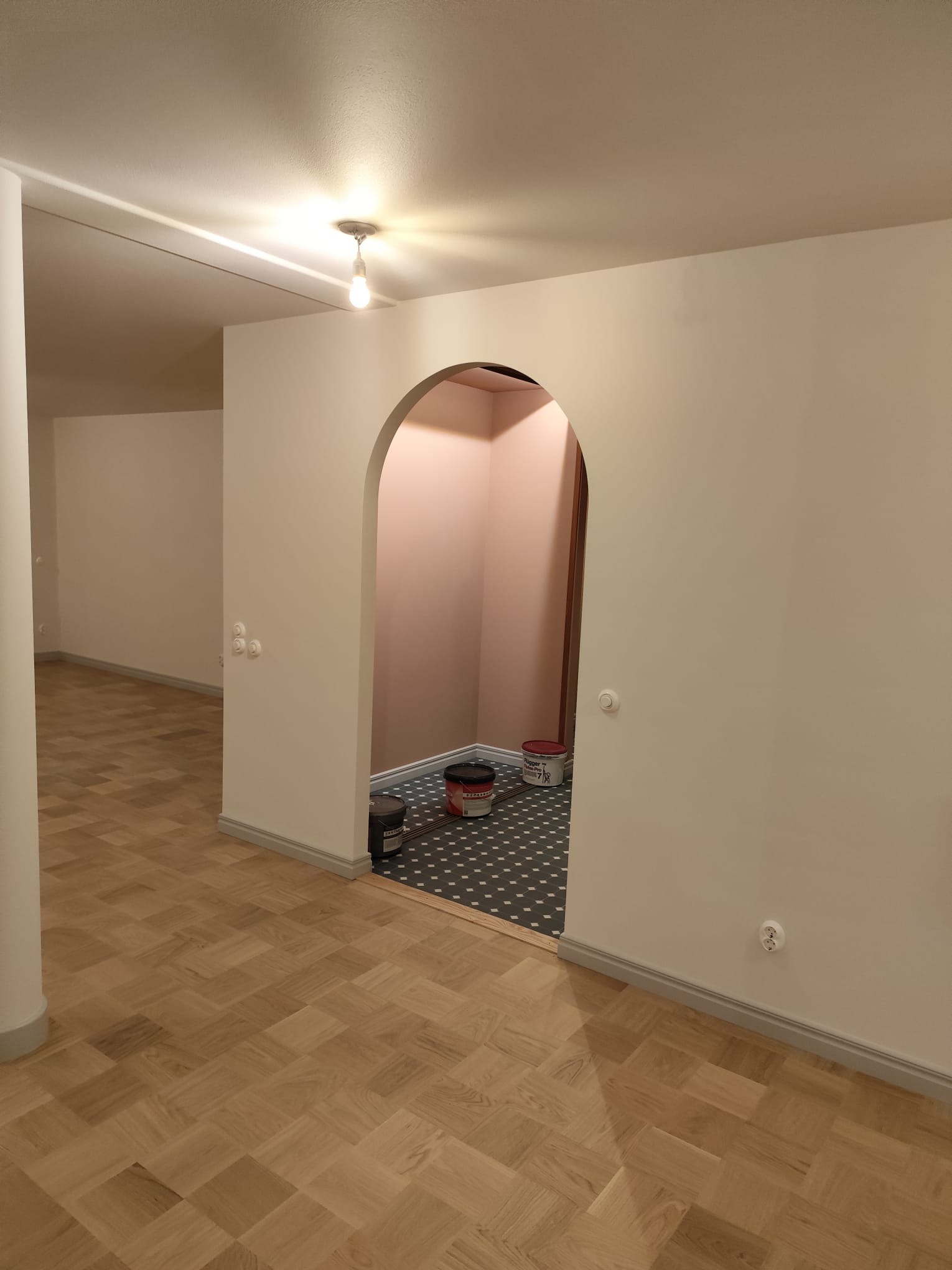 Wnętrze po malowaniu: jasne ściany, parkiet, widoczne puszki farb Flugger w przejściu z łukiem. Świeżo odmalowane pomieszczenie z widocznymi detalami wykończeniowymi.