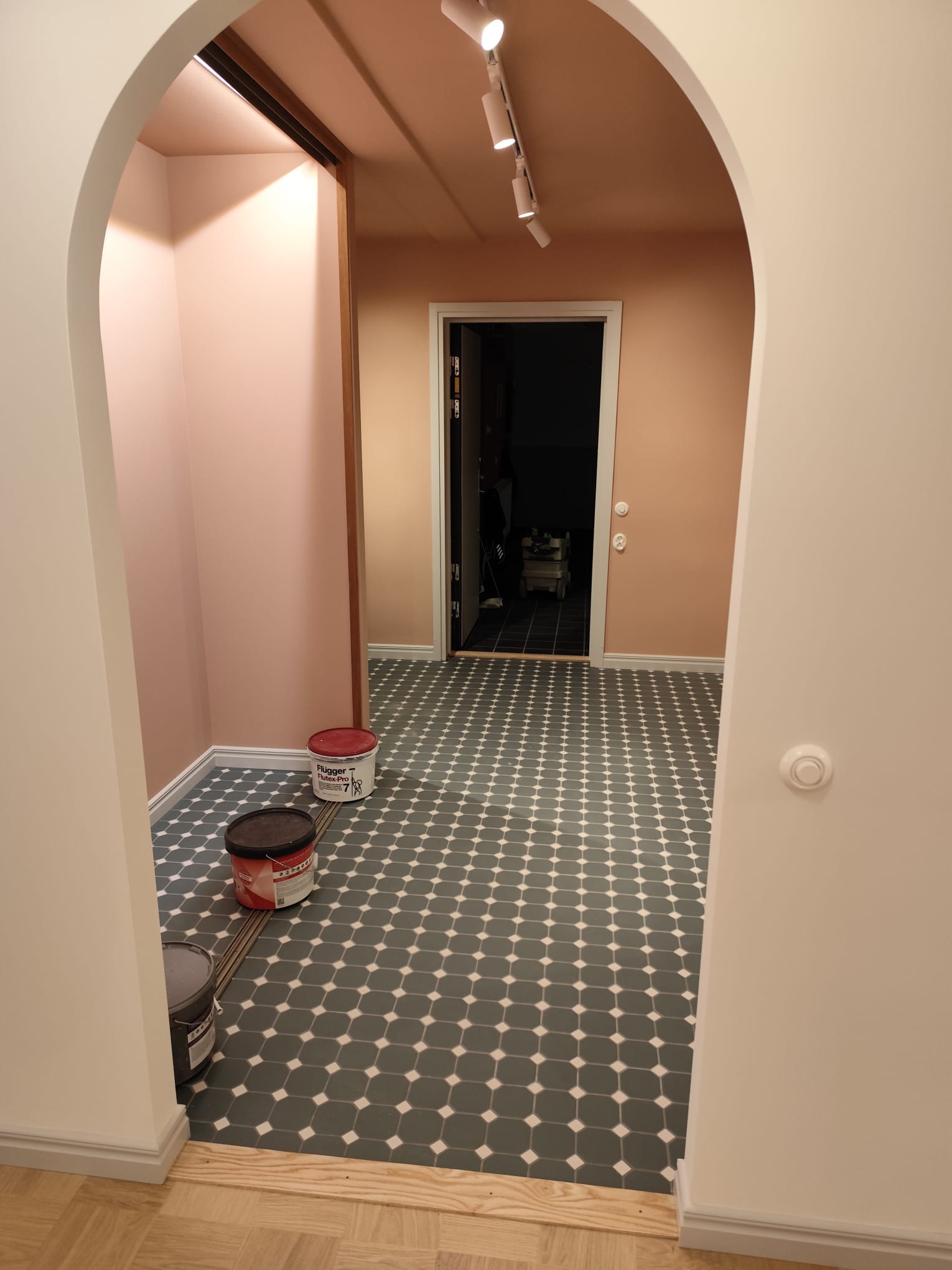 Wnętrze po malowaniu z łukowym przejściem, widoczne puszki z farbą Flugger, szaro-biała podłoga w romby i różowe ściany. Oświetlenie punktowe.