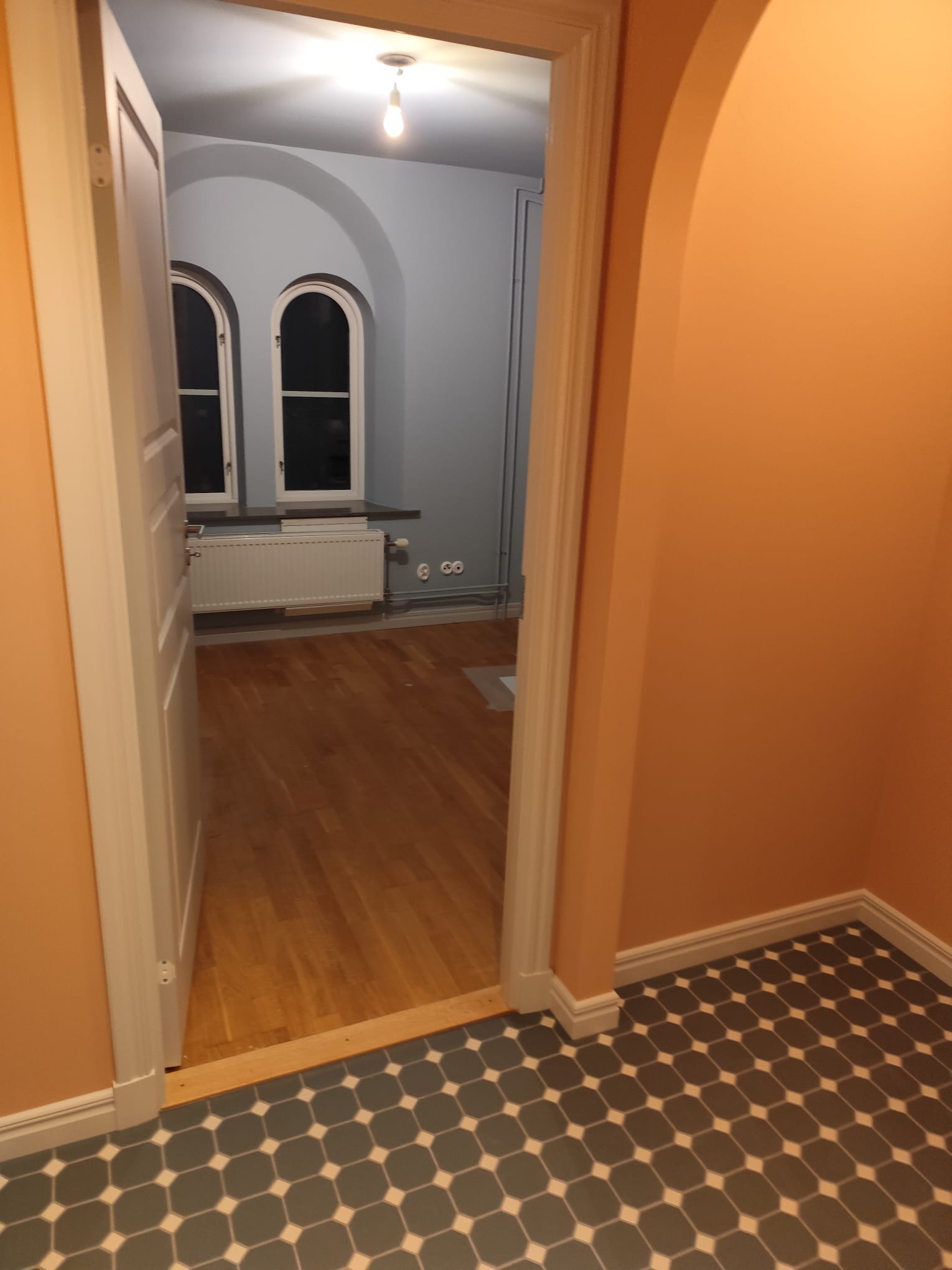 Wnętrze po remoncie: parkiet, pomarańczowe ściany, szara podłoga z wzorem, białe okna łukowe i drzwi. Widok z korytarza do pokoju. Żarówka sufitowa.