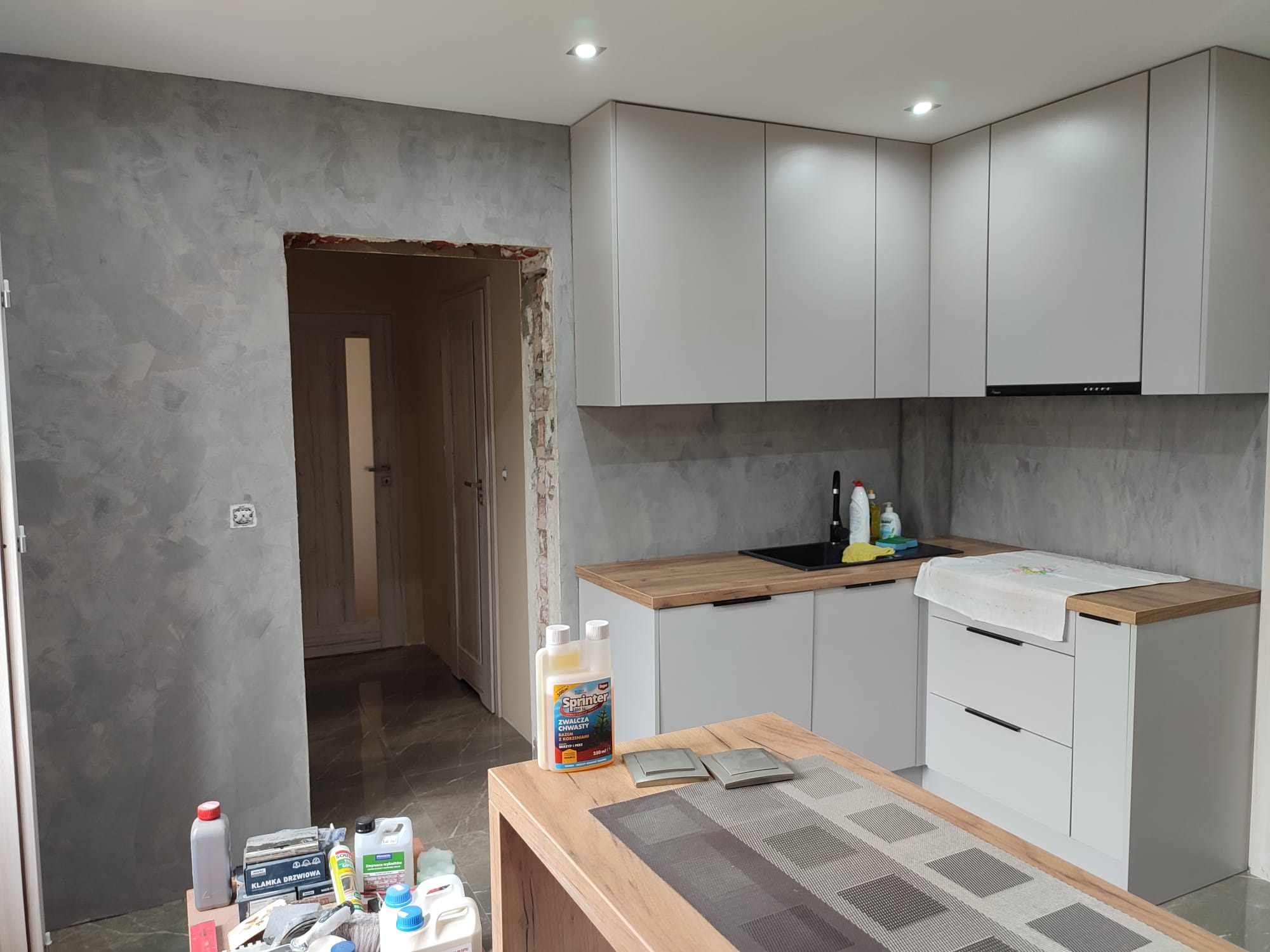 Kuchnia w trakcie wykończenia: szare ściany z efektem dekoracyjnym, białe szafki kuchenne z drewnianym blatem, widoczne narzędzia i materiały malarskie, surowy otwór drzwiowy.