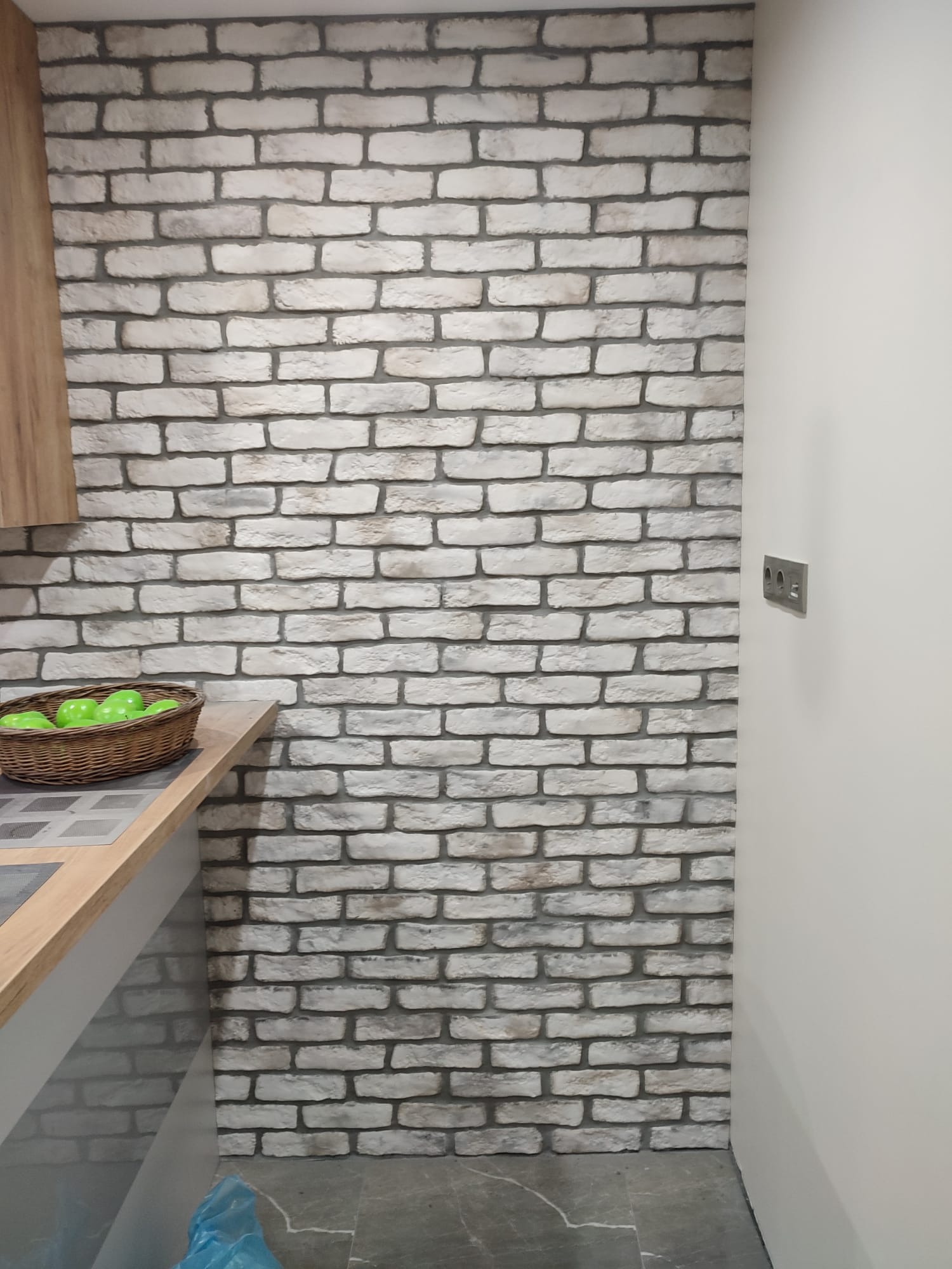 Ściana wykończona dekoracyjną cegłą w odcieniach bieli i szarości, widoczny fragment blatu kuchennego z koszykiem zielonych jabłek i gładka, biała ściana z gniazdkiem elektrycznym.